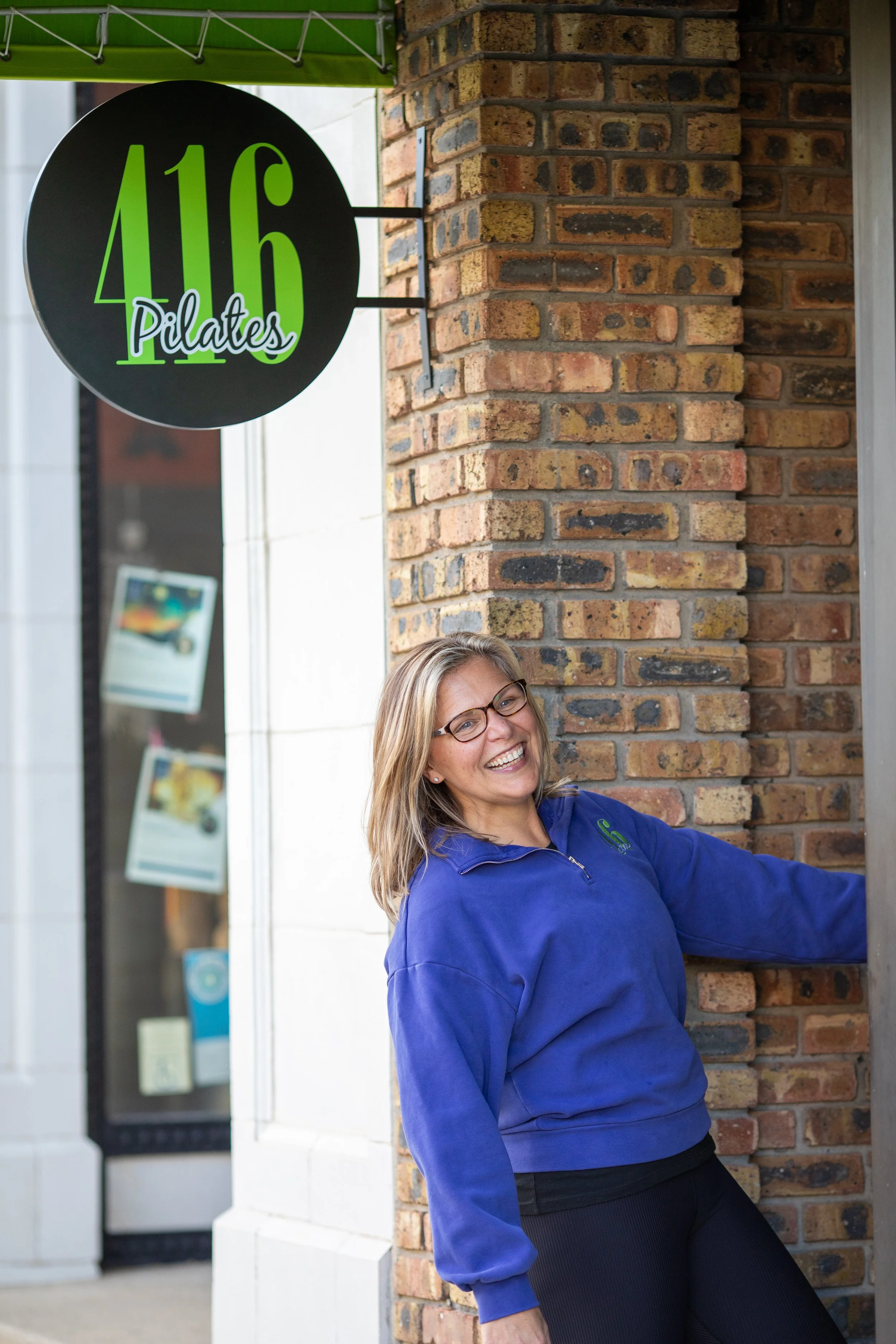 A woman smiling and leaning against a brick wall near a sign that reads '416 Pilates'