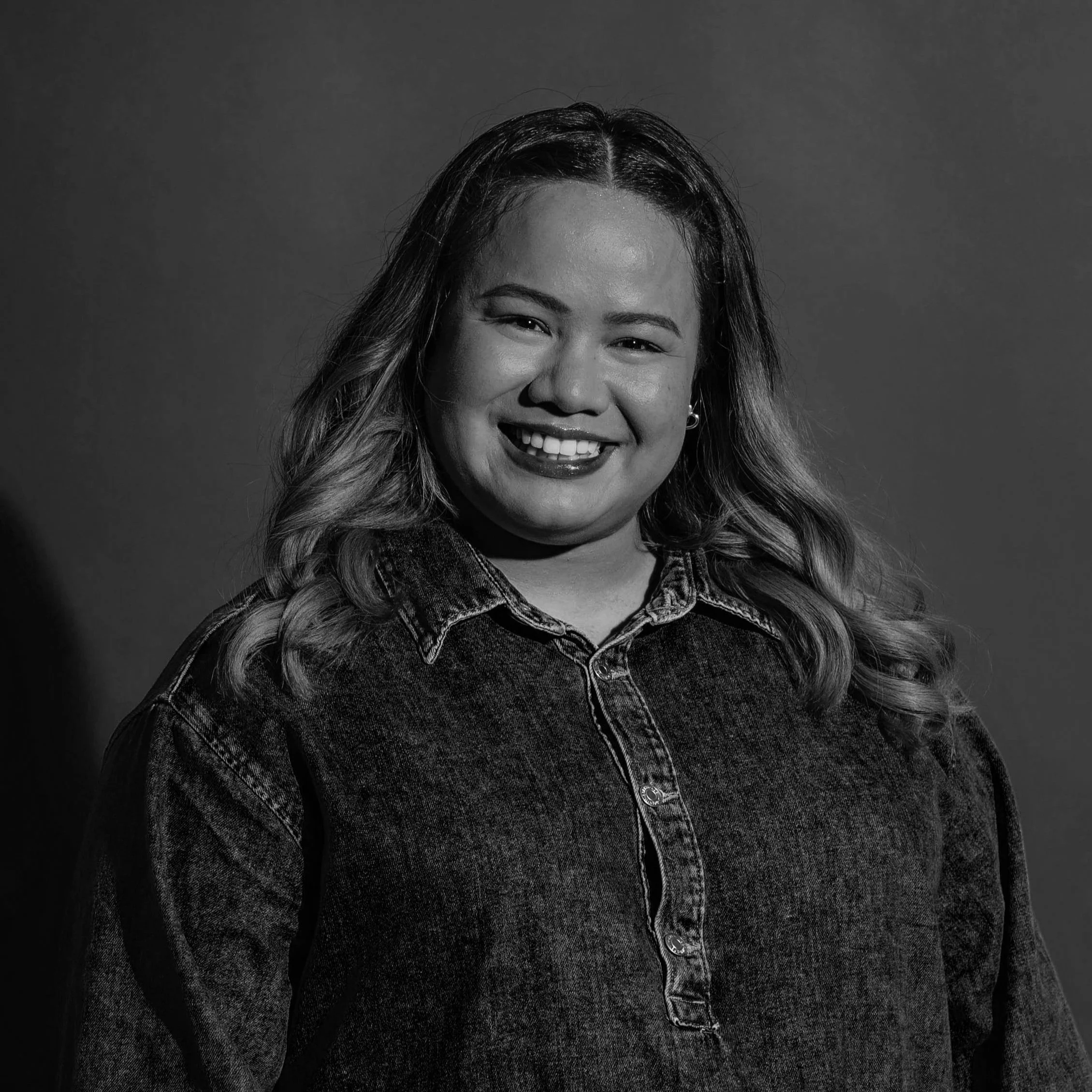Black and white portrait of a woman with long wavy hair, smiling, wearing a denim shirt, against a plain background.