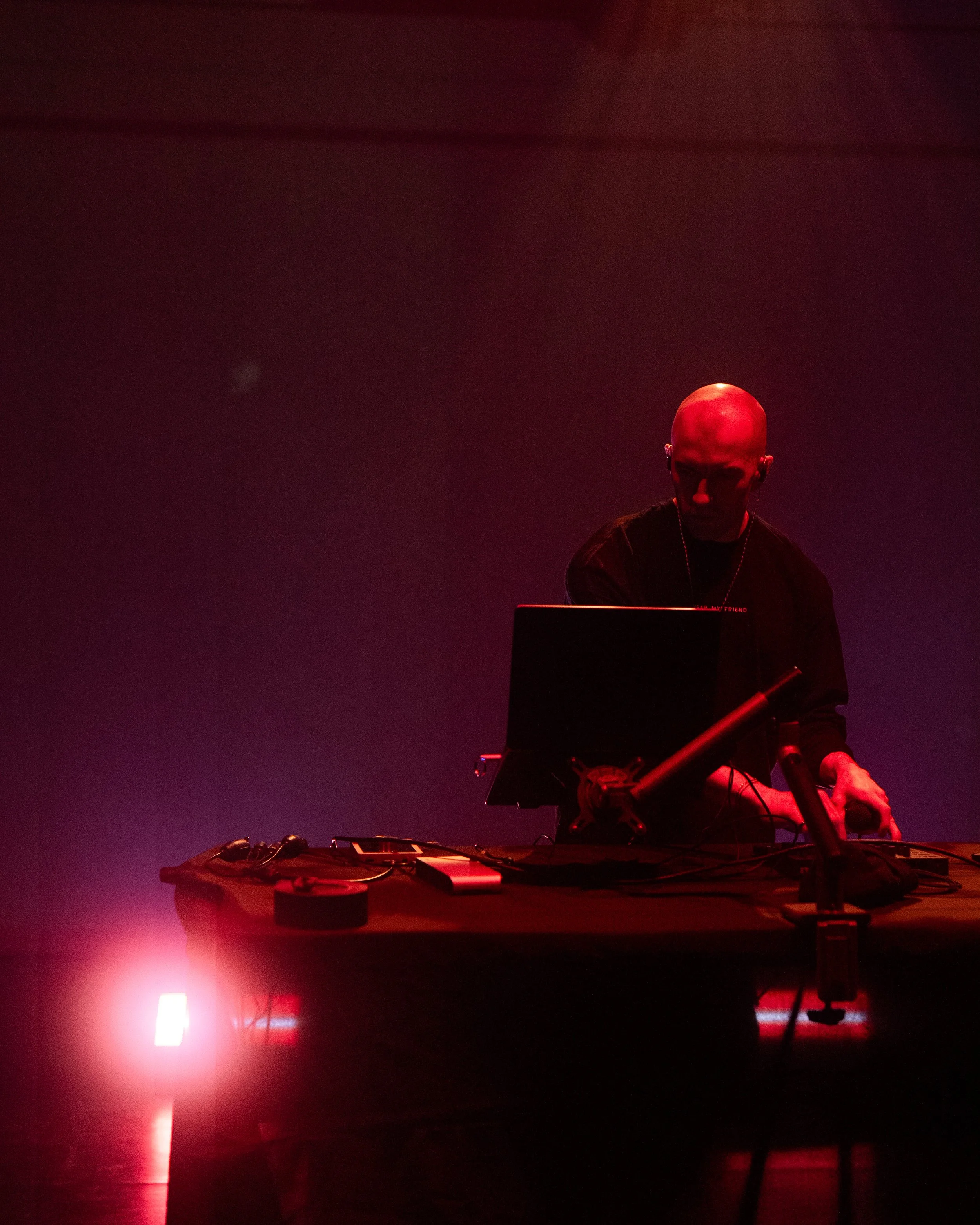 A solitary bald person performing or working DJ setup with red stage lighting, a laptop, and electronic equipment on a table.