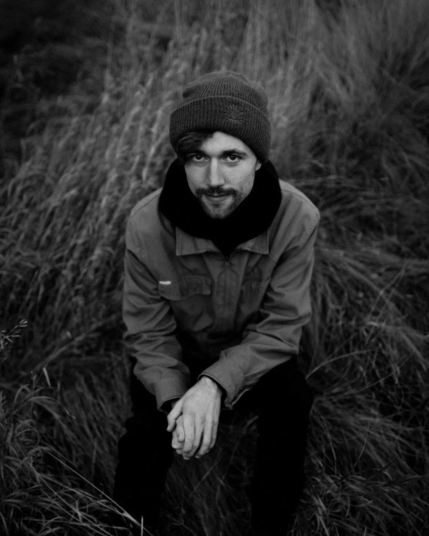 A young man with a beard and mustache wearing a beanie, jacket, and scarf, sitting on the ground in a field of tall grass, looking up at the camera.