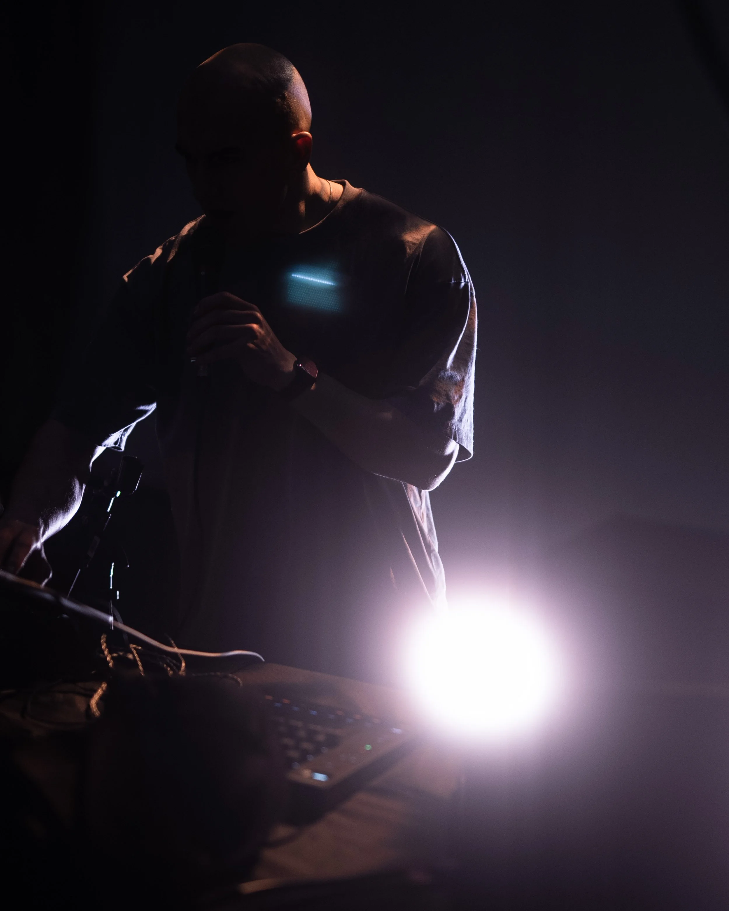 Silhouette of a person with a shaved head working with DJ equipment in low light, illuminated by a bright light source.