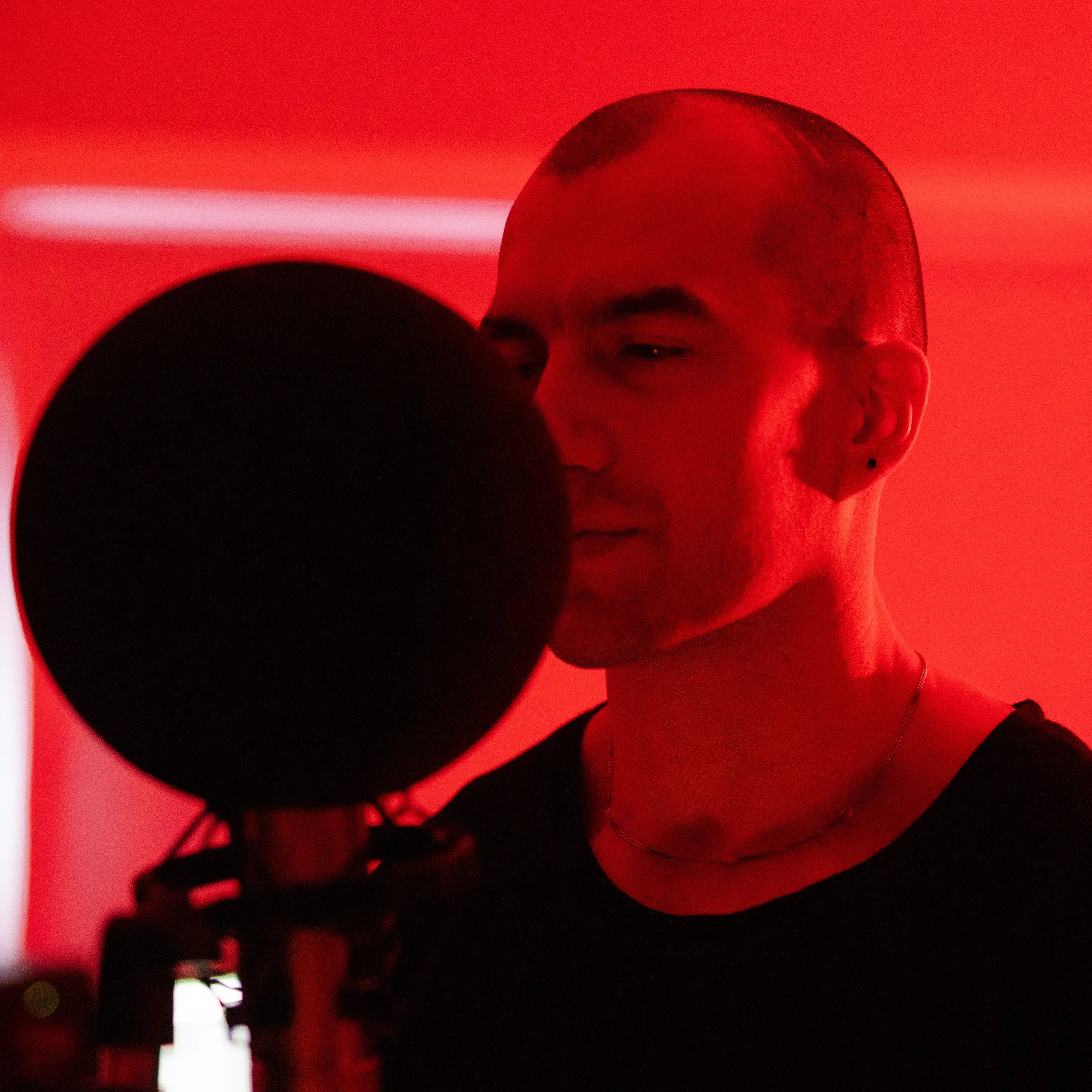 A person with a shaved head and earring recording vocals in a red-lit studio with a microphone.