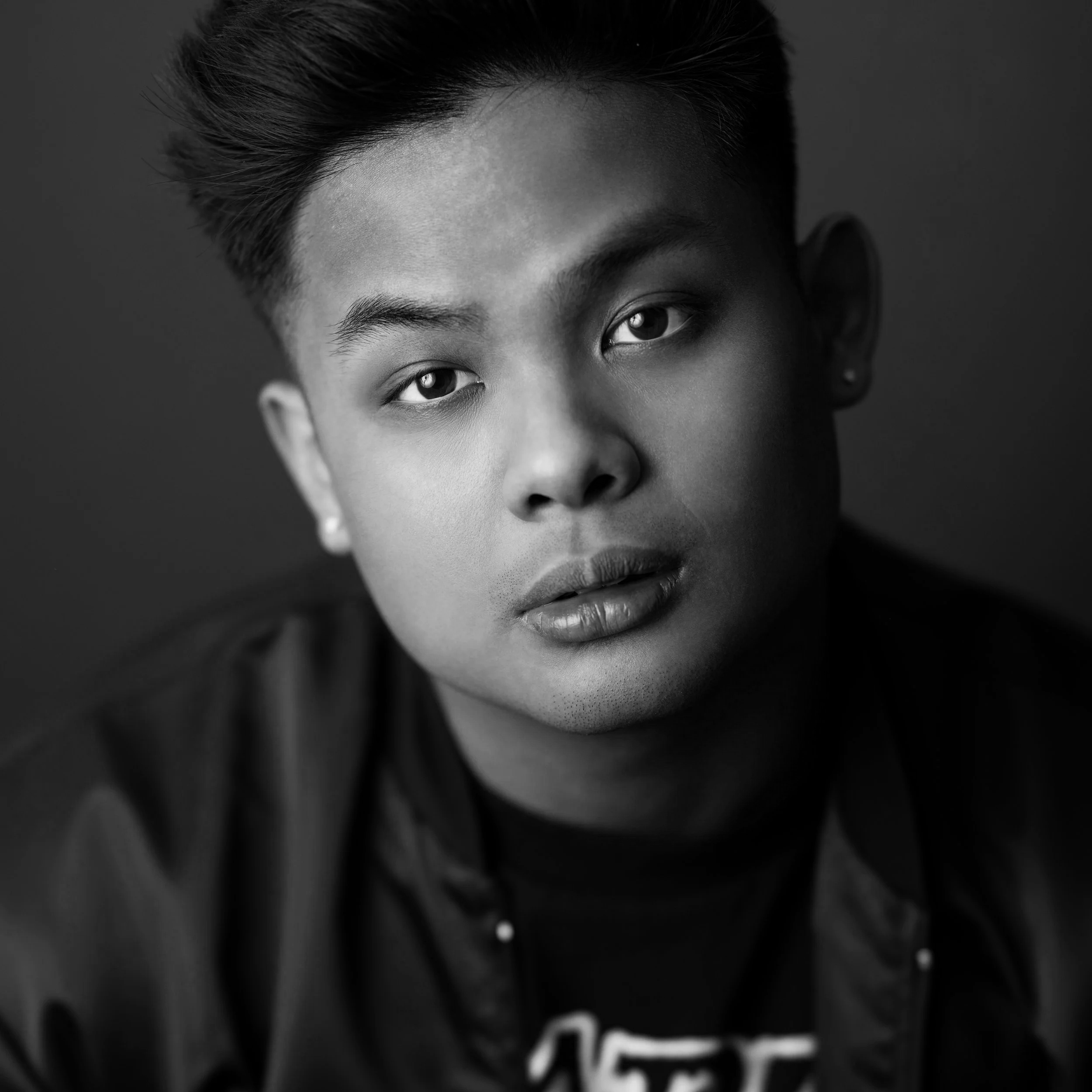 Black and white portrait of a young person with short hair, wearing earrings and a dark jacket, looking directly at the camera.