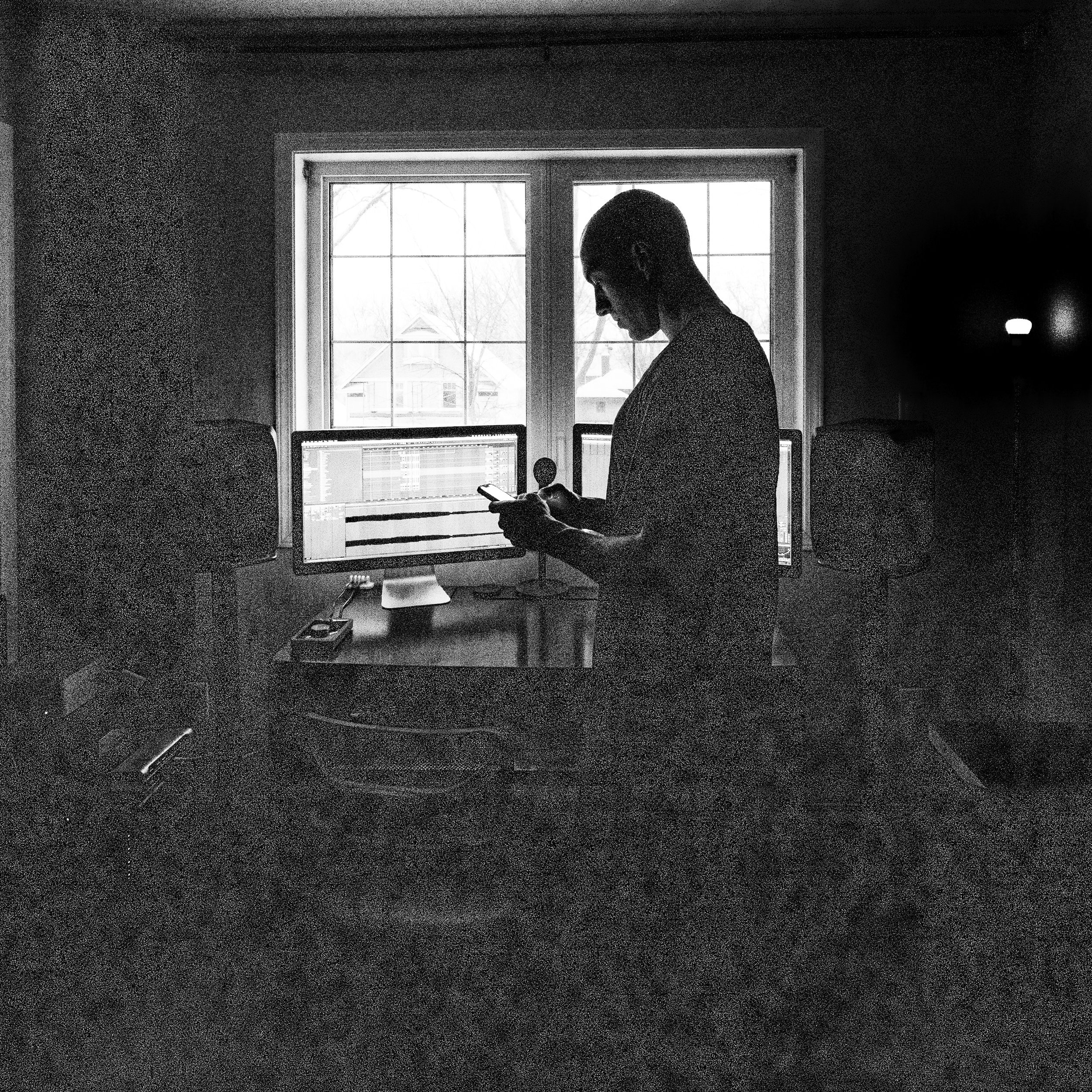 A person standing in front of a desk with two computer monitors, looking at a phone, with a window in the background showing a house and trees. The room is dimly lit.
