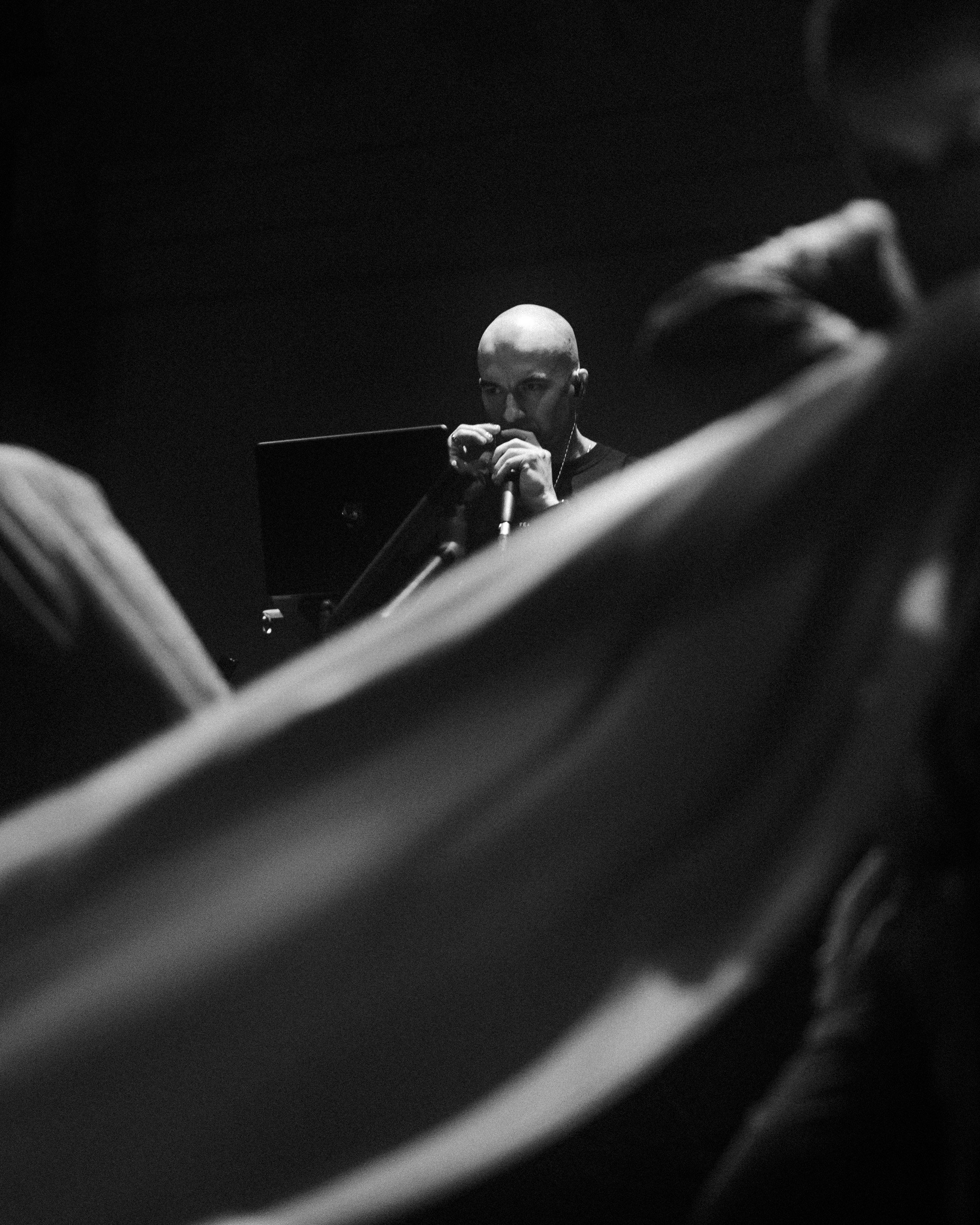 A black and white photo of a person with a bald head, wearing headphones, holding a microphone, and standing in front of a monitor, seen through blurry foreground objects.