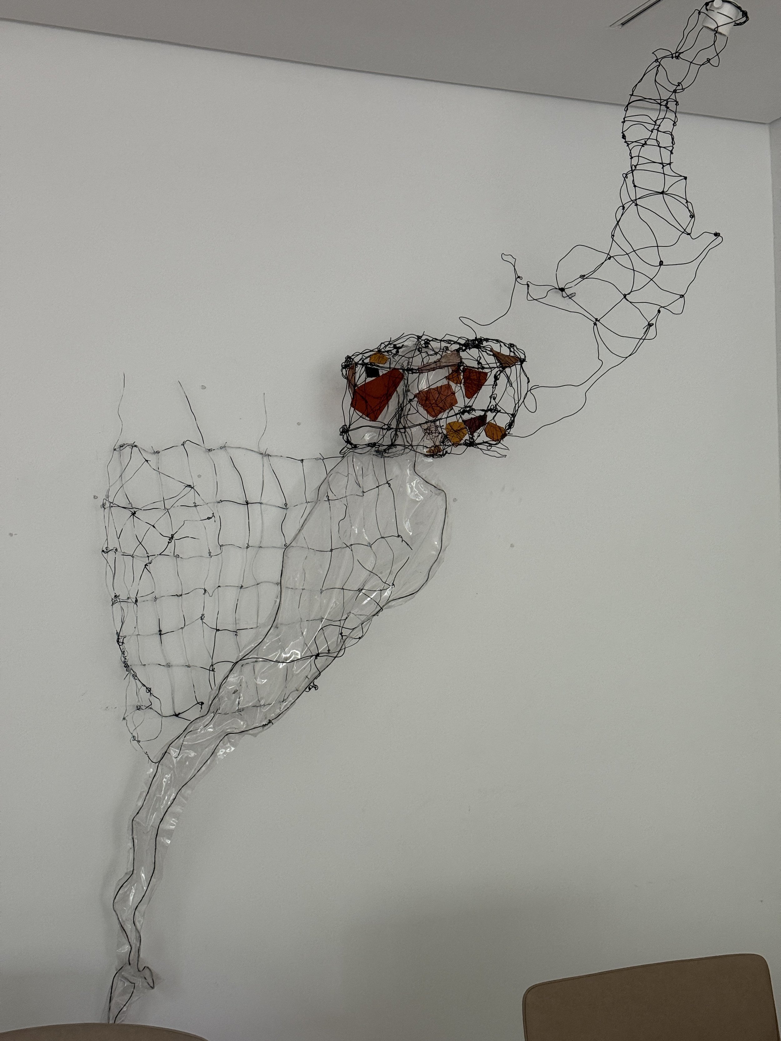 Metal wire sculpture with colored glass pieces, mounted on a wall, with some wire extending downward.