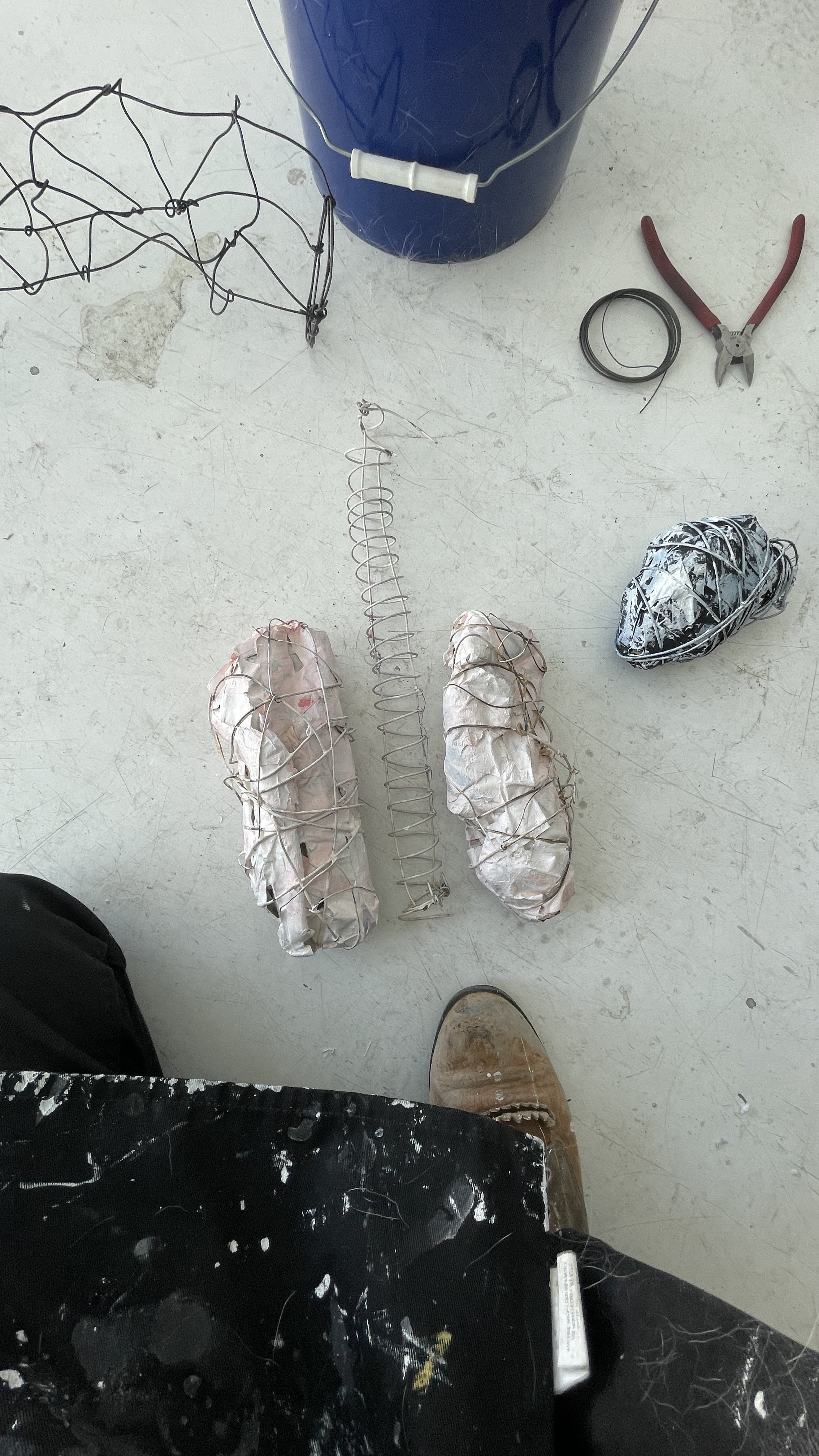 Photo of a workspace with various DIY project materials, including a wire frame structure, foam-wrapped objects, a coil spring, a pair of pliers, and a black bag with paint splatters. A person's brown boots and black pants are visible at the bottom o