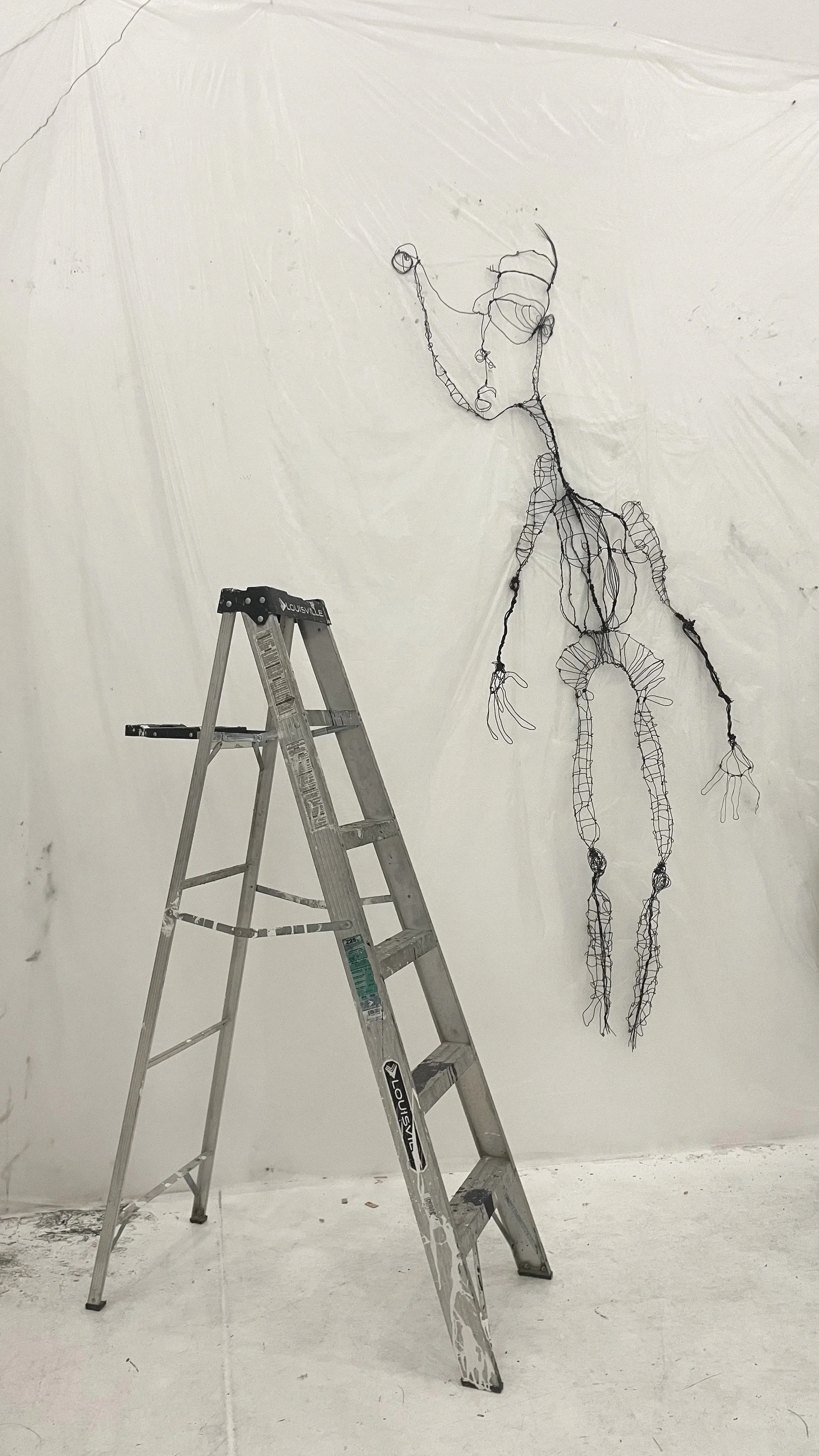 Wire sculpture of a human figure on a blank white wall, with a ladder nearby.