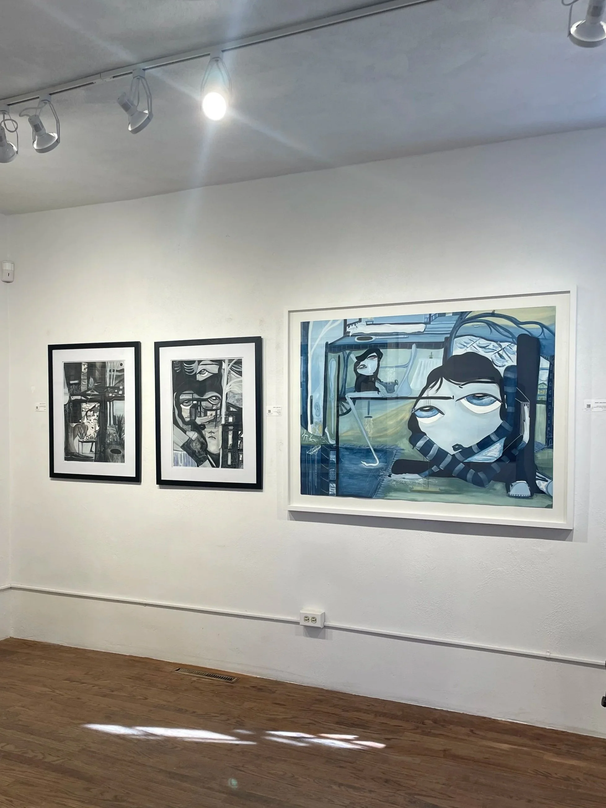 Three paintings on a white gallery wall, including two black and white and one blue-themed artwork, with wooden floor and ceiling lighting track.