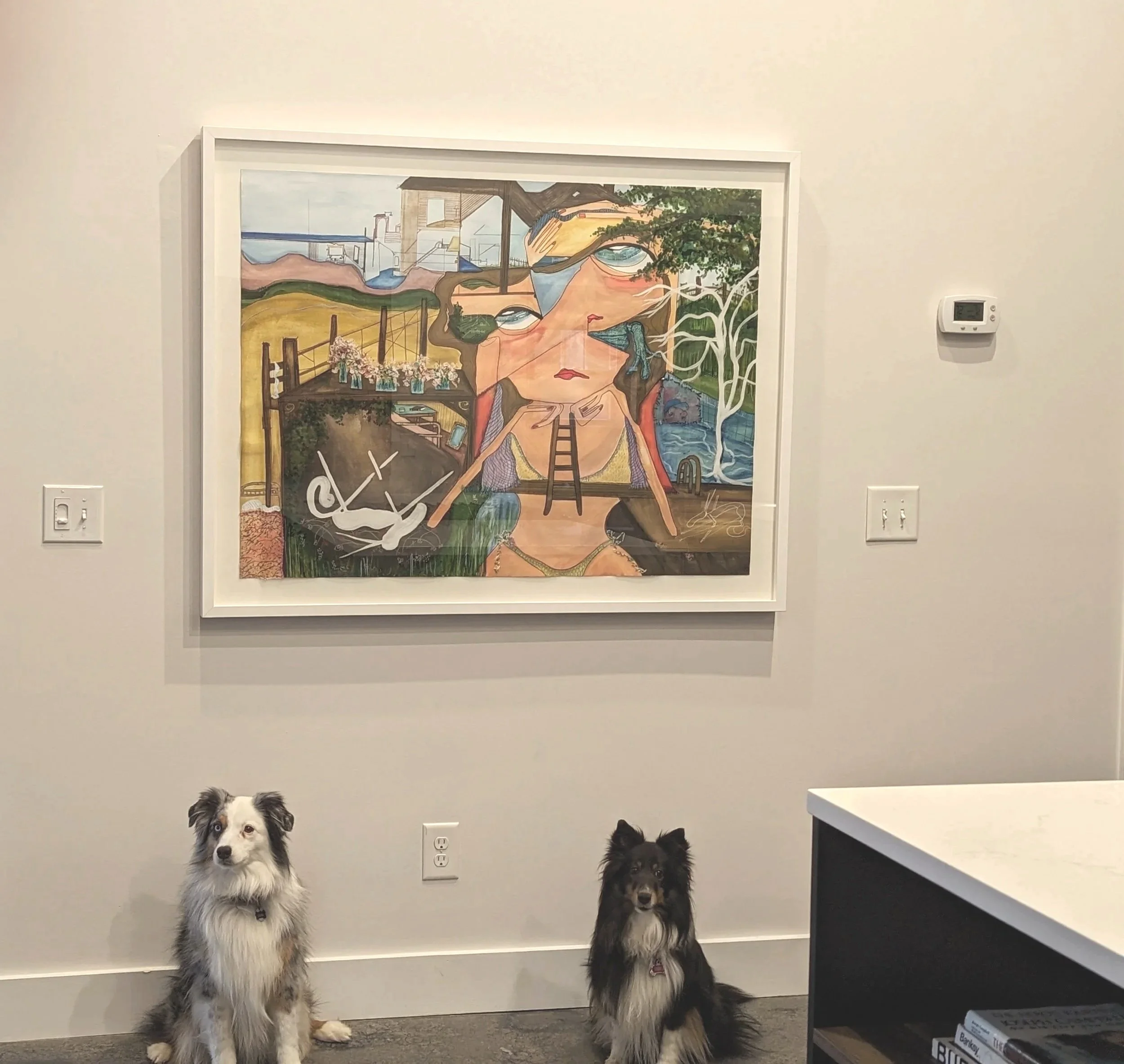 Two Border Collies sitting on the floor in front of a wall with a colorful abstract painting. The painting features a large human face, a ladder, trees, water, and various abstract elements.