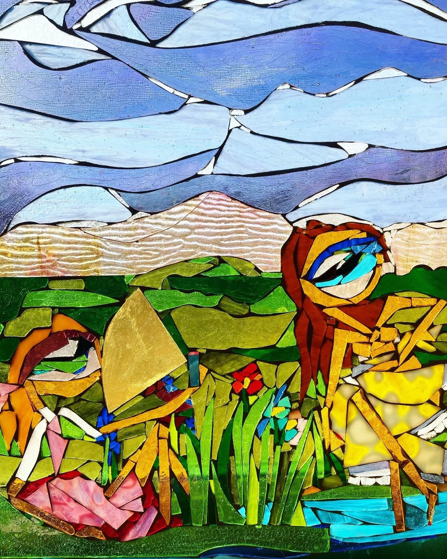 A mosaic stained glass art depicting mountains, a sunset, green hills, and a woman with red hair sitting by a yellow flower and some tall green grass.
