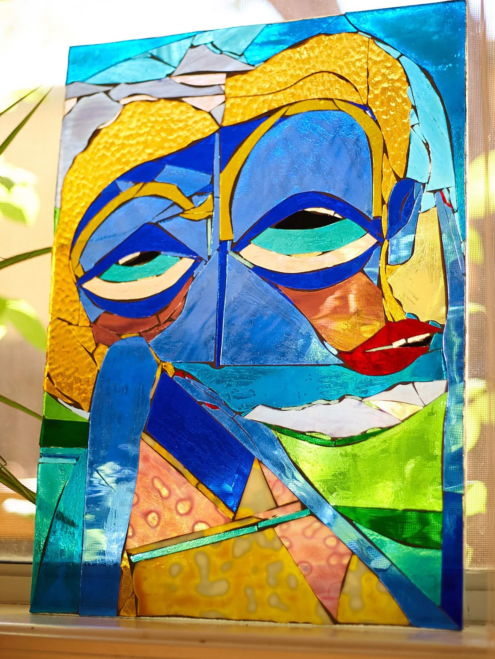 Colorful stained glass art portraying a person's face with blue skin, yellow hair, and red lips, resting their head on their hand.