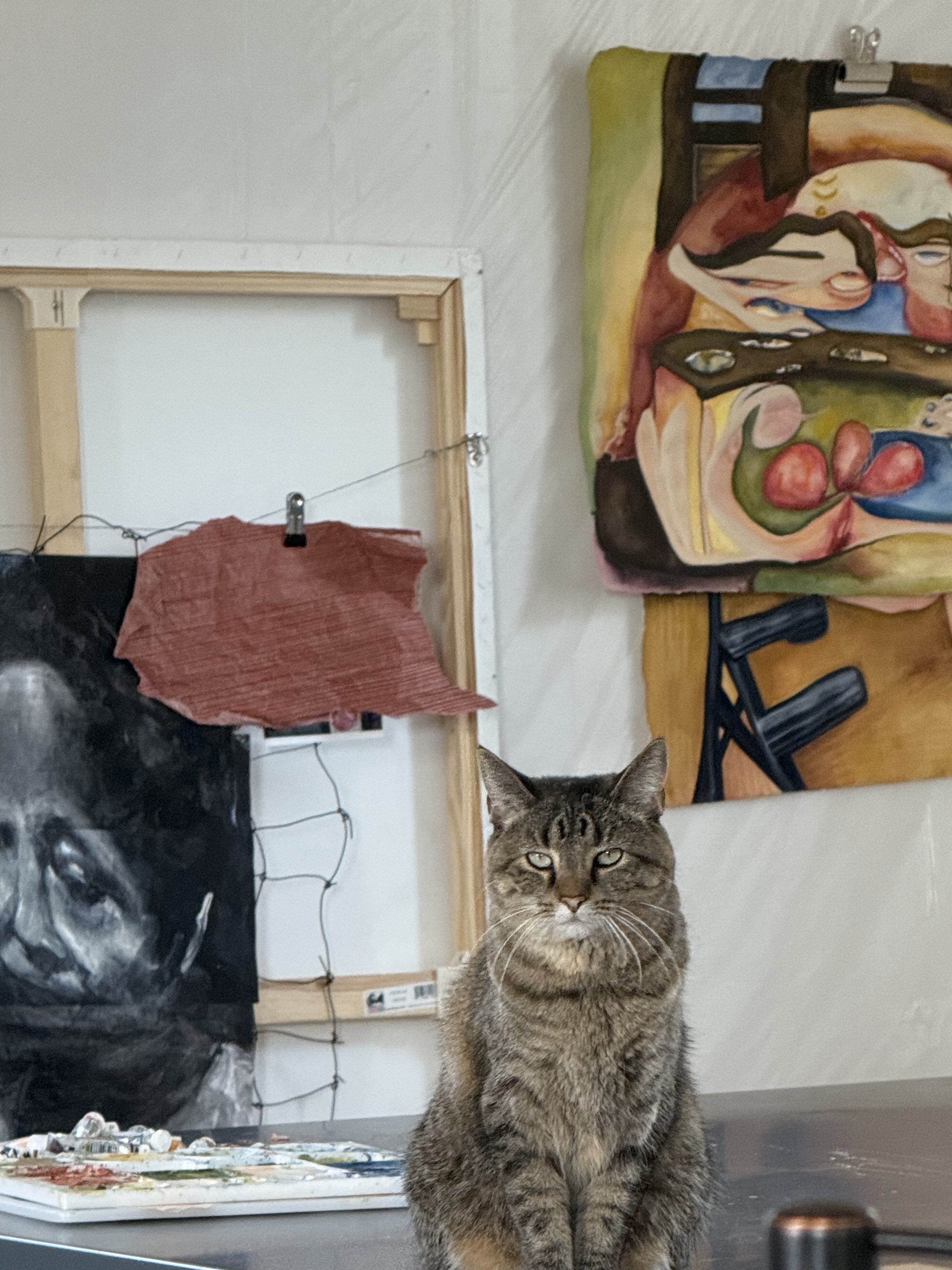 A tabby cat sitting on a table with an art palette and paints, looking directly at the camera, with a painting and various objects in the background.