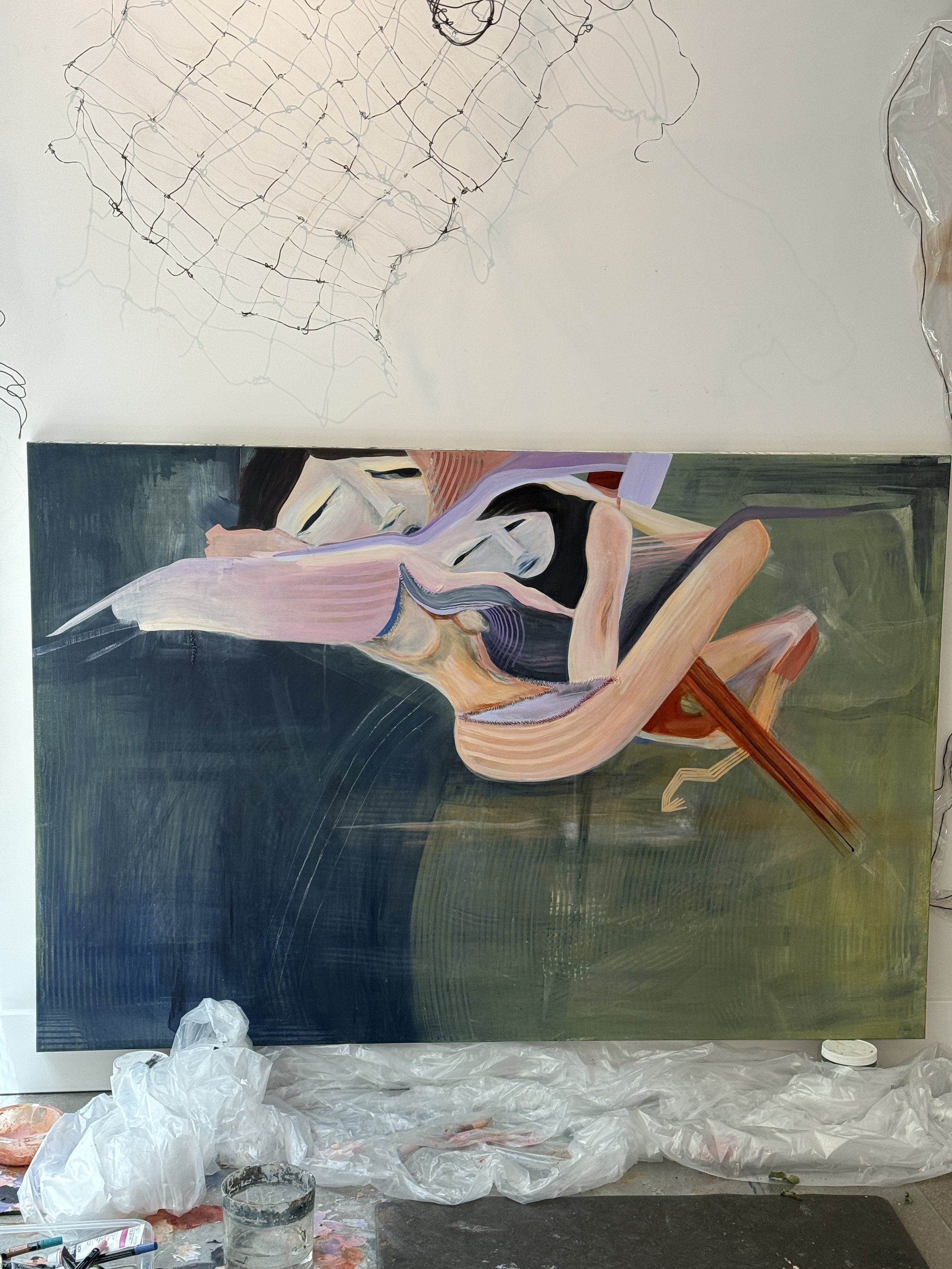 A painting in progress on a large canvas depicts an abstract image of a woman with dark hair and a black top. The artist's workspace includes plastic sheets, brushes, and containers of paint on the floor.