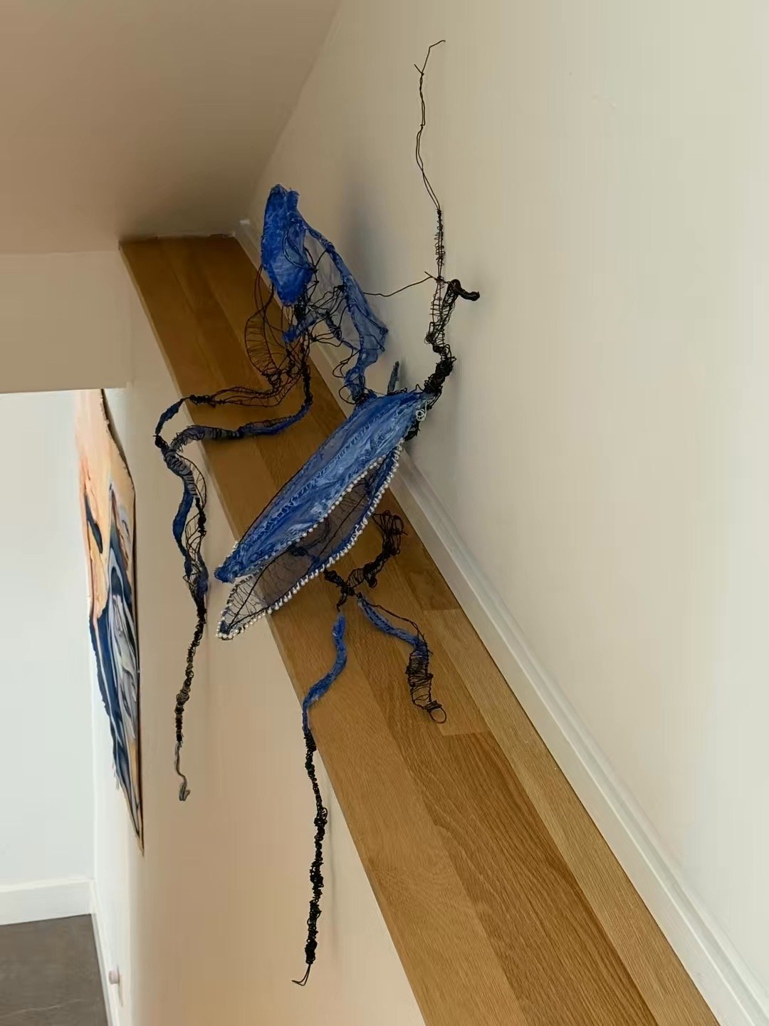 Contemporary art sculpture made of tangled black and blue wire, mounted on a white wall in a room with wooden flooring.