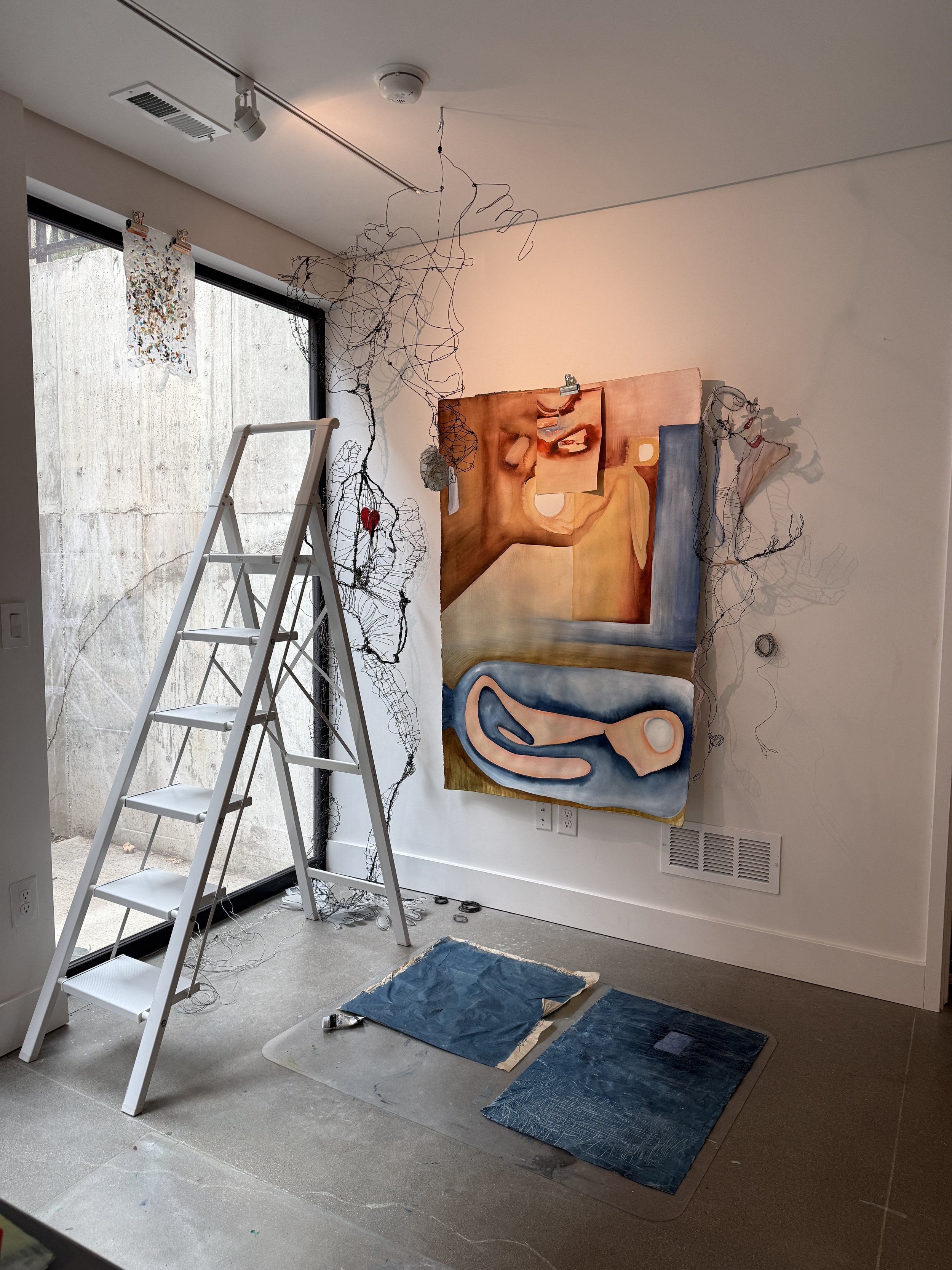 An indoor art studio with a large abstract painting on the wall, a stepladder, and various painting supplies on the floor, including canvases and protective sheets.