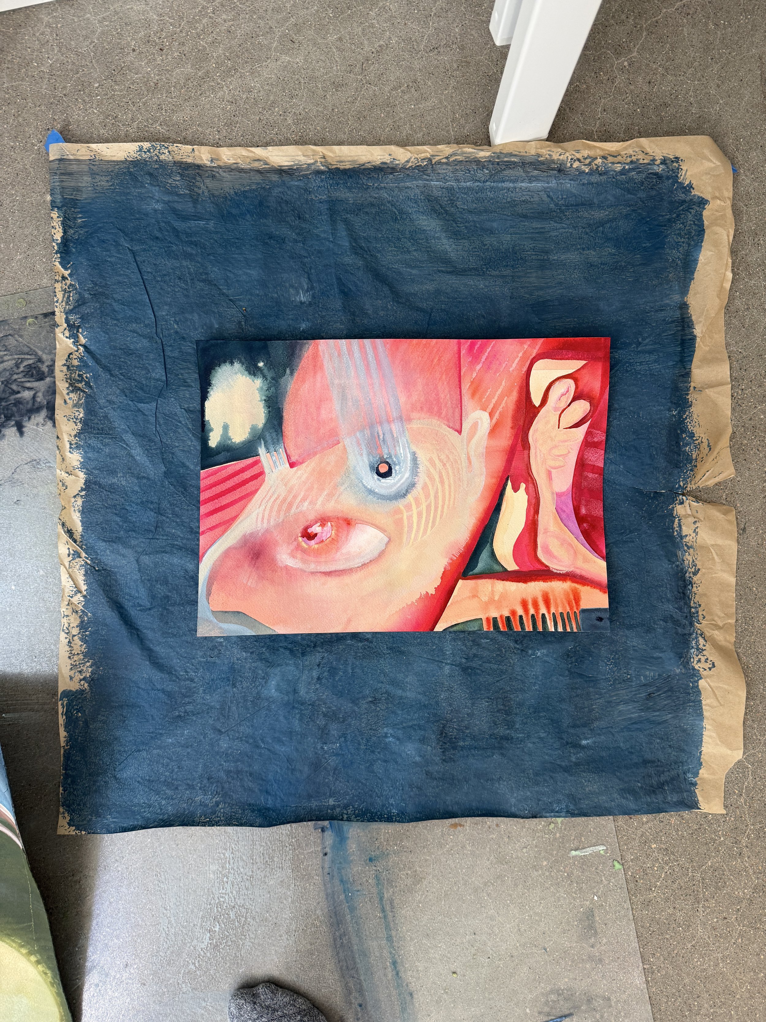 A colorful painting of a person's face with blonde hair, pink lips, and a yellow background, lying on a black surface that appears to be painted with blue and black paints, next to a white table leg.