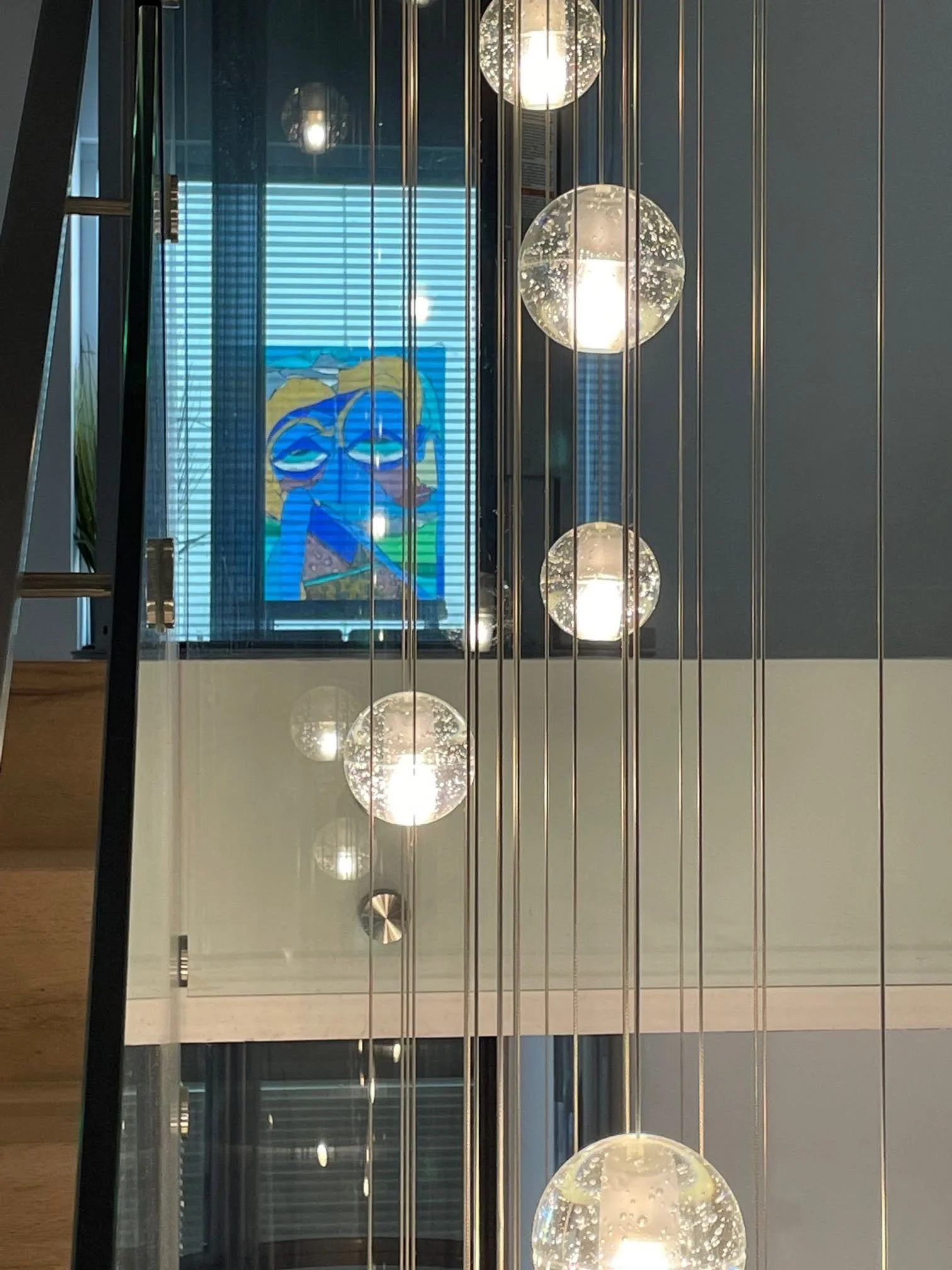 Interior staircase with a glass railing, modern spherical pendant lights hanging vertically, and a colorful abstract painting on a window in the background.
