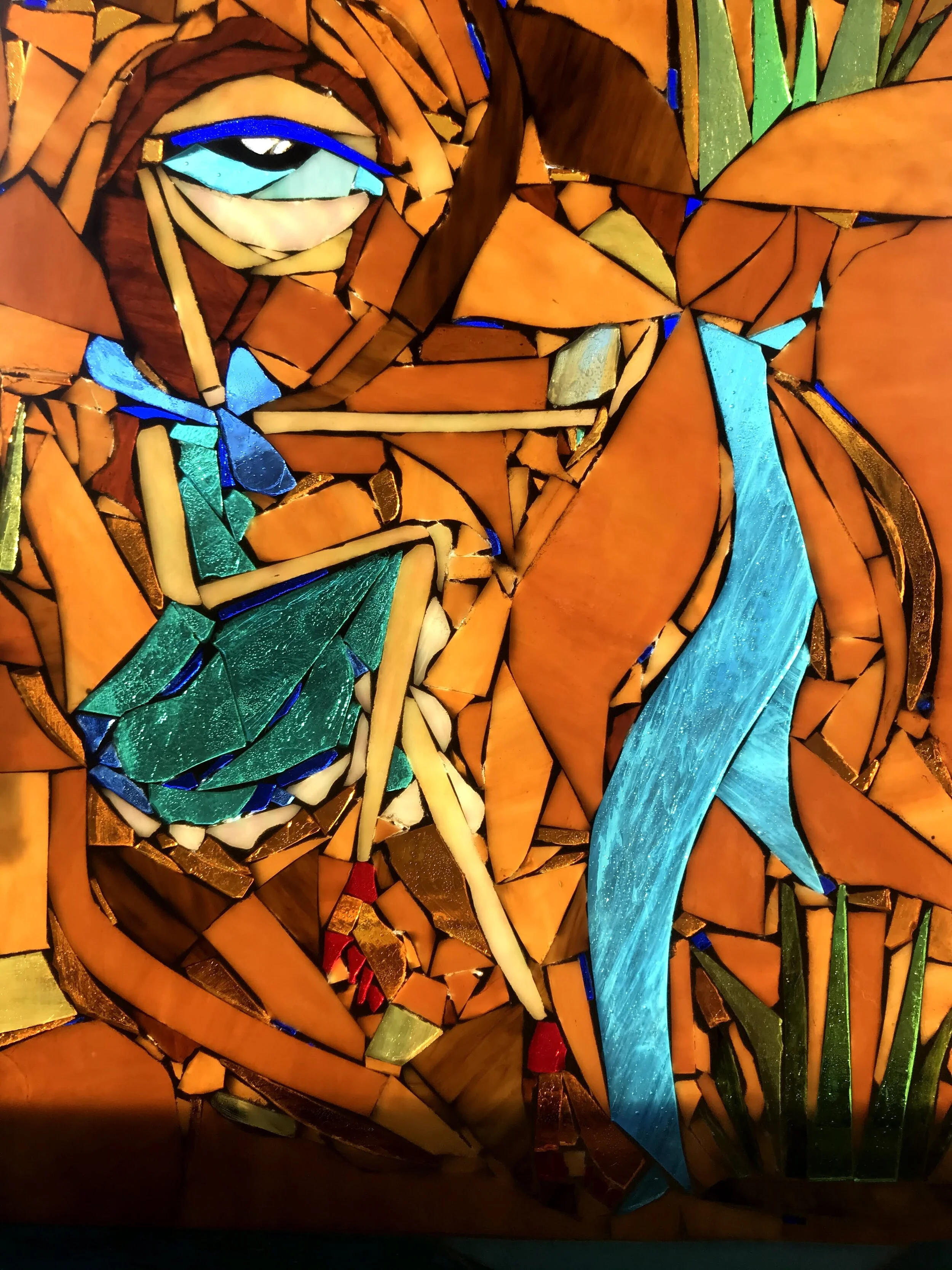 A stained glass artwork depicting a woman holding a fishing pole, with orange, blue, green, and beige glass pieces arranged to form the scene.