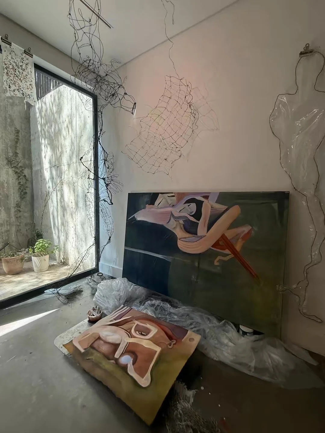 An art studio with a large painting of a woman sitting on a wooden chair. The studio contains two other unframed paintings, wire sculptures hanging from the ceiling, and a window with sunlight coming through. Potted plants are near the window.