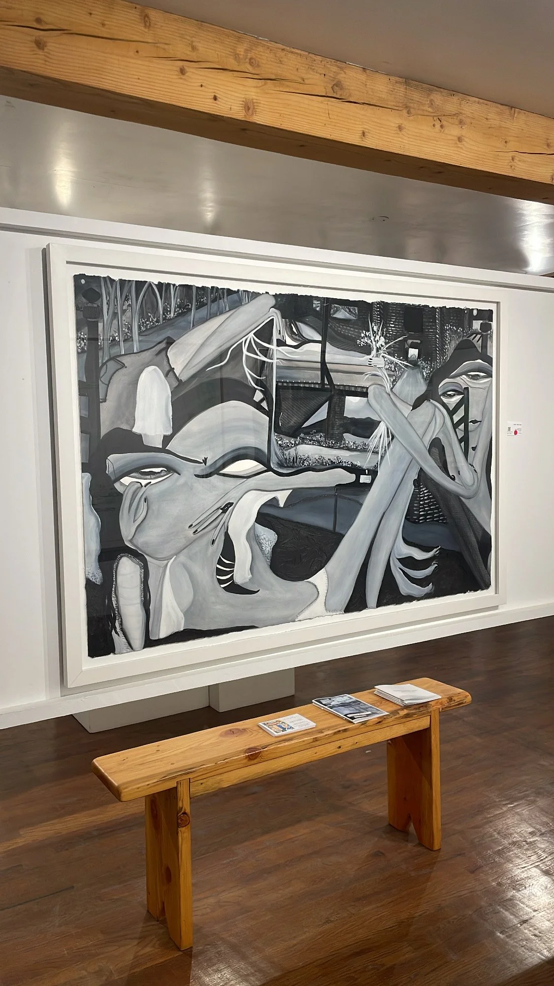 A large black and white abstract painting featuring distorted human faces and forms, displayed on a gallery wall with a wooden bench and small brochures in front.