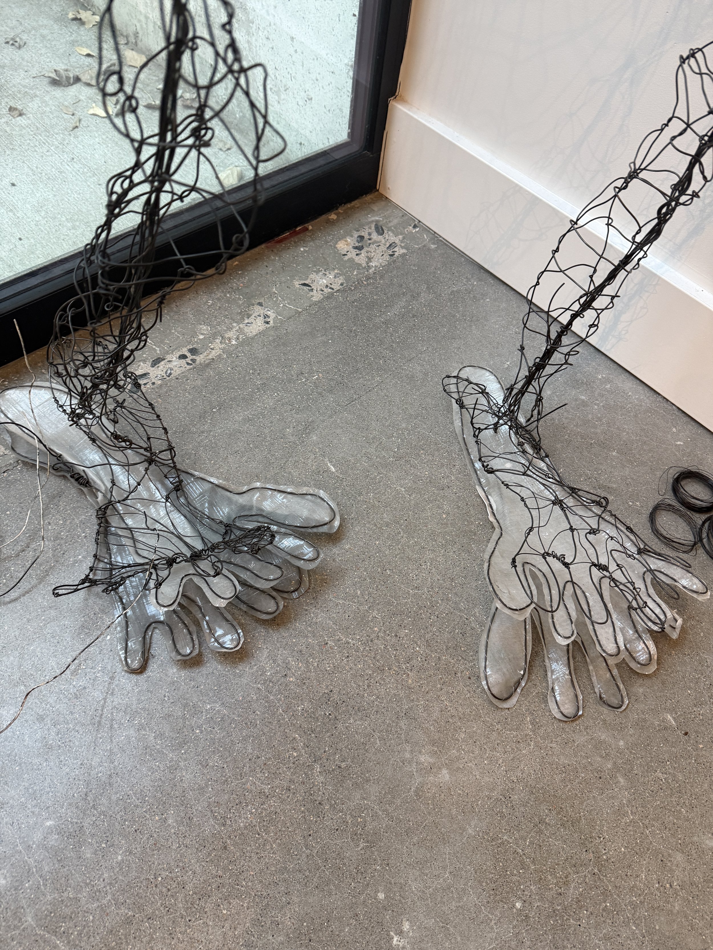 Close-up of a pair of wireframe angel wings with clear plastic bases, placed on a concrete floor near a glass door.
