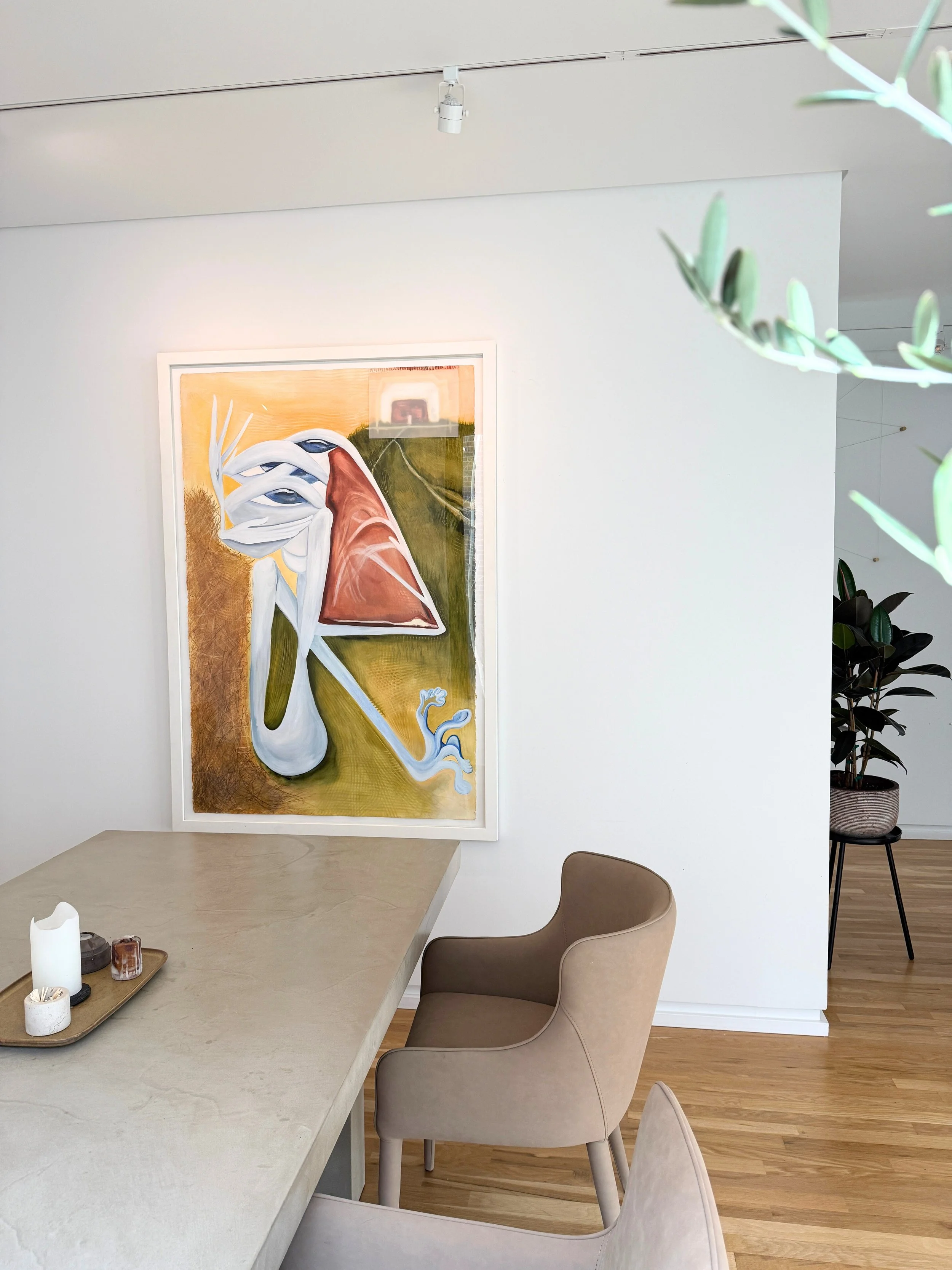 Interior of a modern dining room with a large abstract artwork on the wall, beige upholstered chair, and a table with candles on a tray, wooden flooring, and a potted plant in the corner.