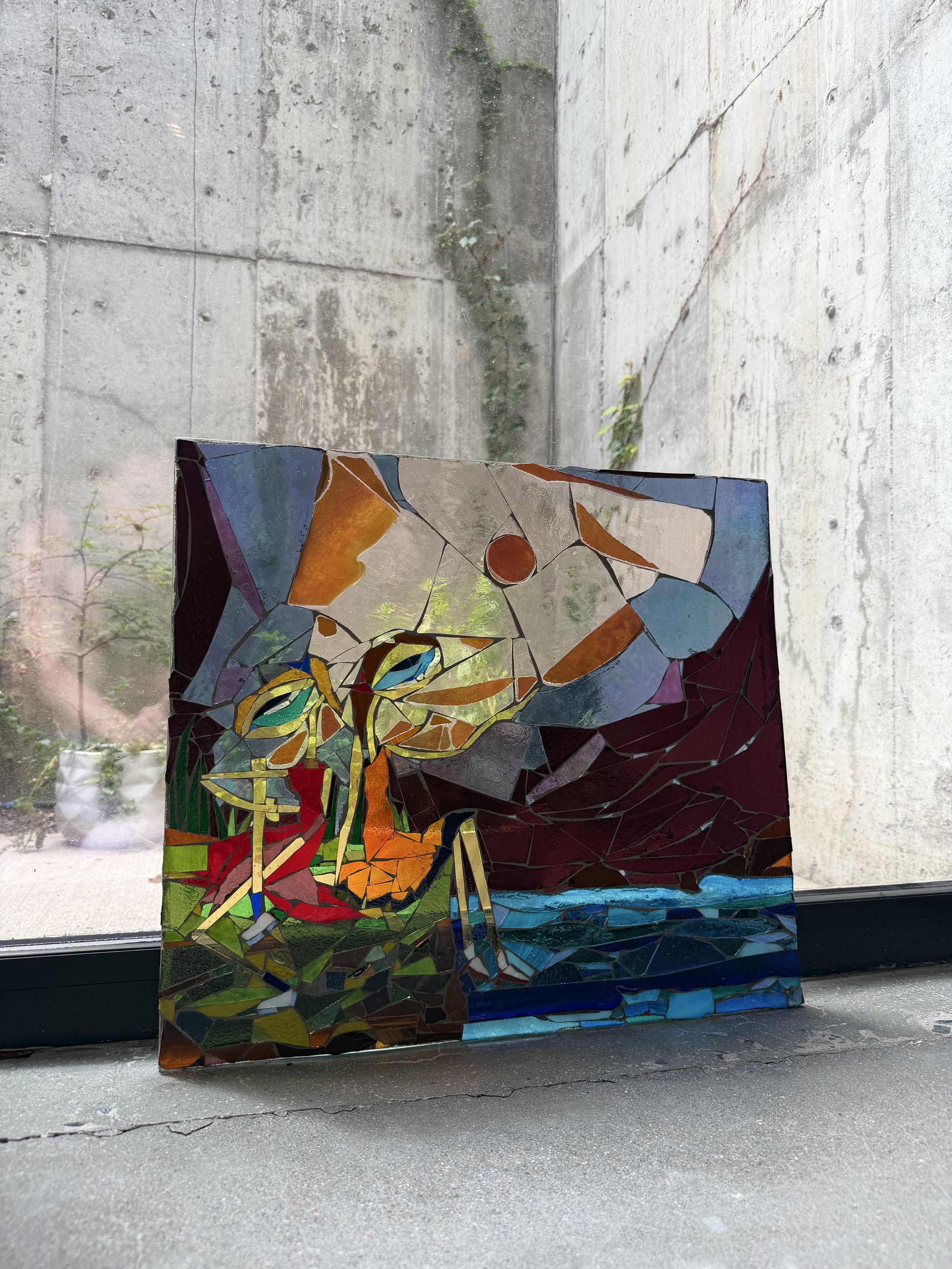 Colorful mosaic butterfly artwork on a rectangular piece of glass in front of a concrete wall and windows with moss and plants outside.