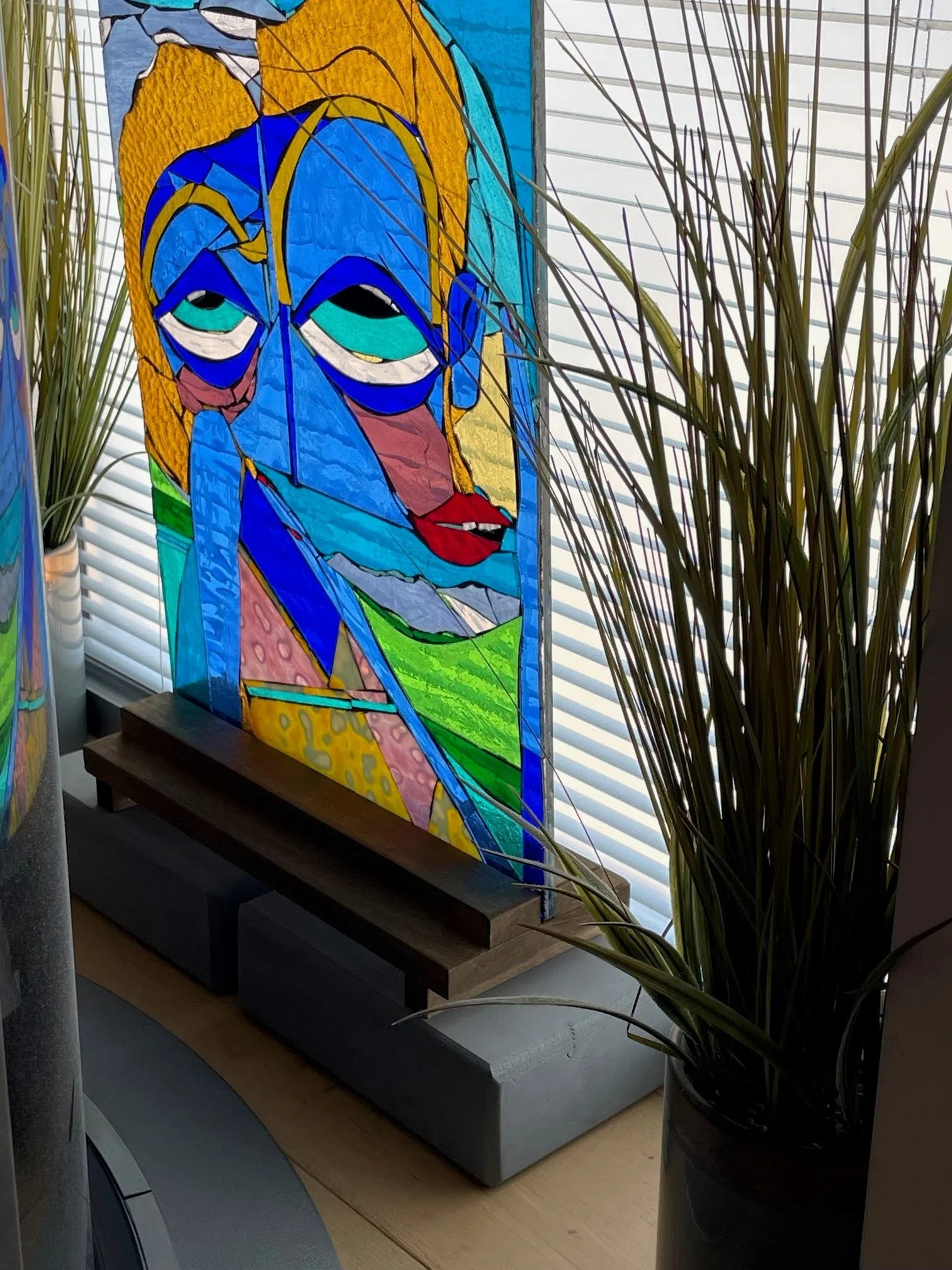 Colorful stained glass window of a woman's face with blonde hair, blue eyes, and red lips, partially obscured by surrounding plants.