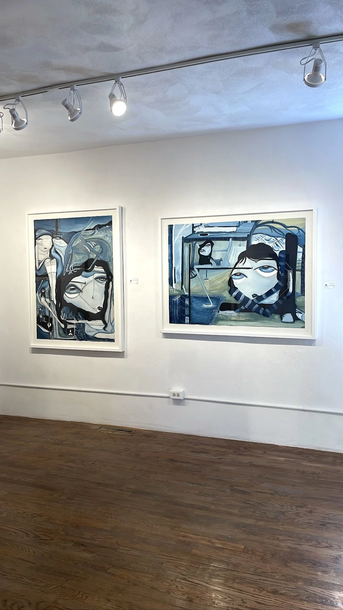 Two abstract paintings with faces hang on a white gallery wall, with track lighting illuminating them.