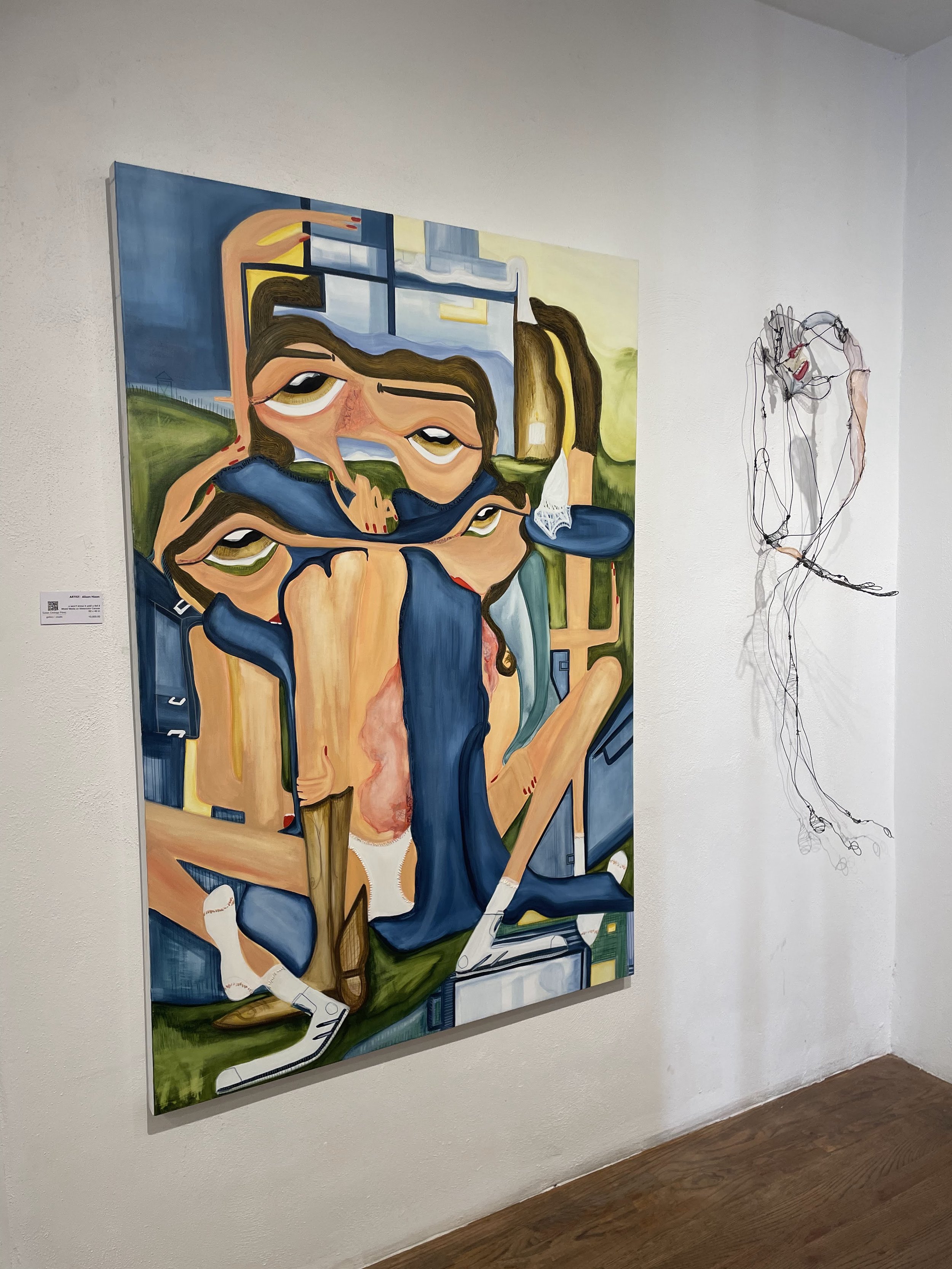 A colorful, abstract painting of a woman with long hair sitting at a desk, with her head resting on her hand and a laptop in front of her, in an art gallery. Next to the painting is a wire sculpture of a head with red lips.
