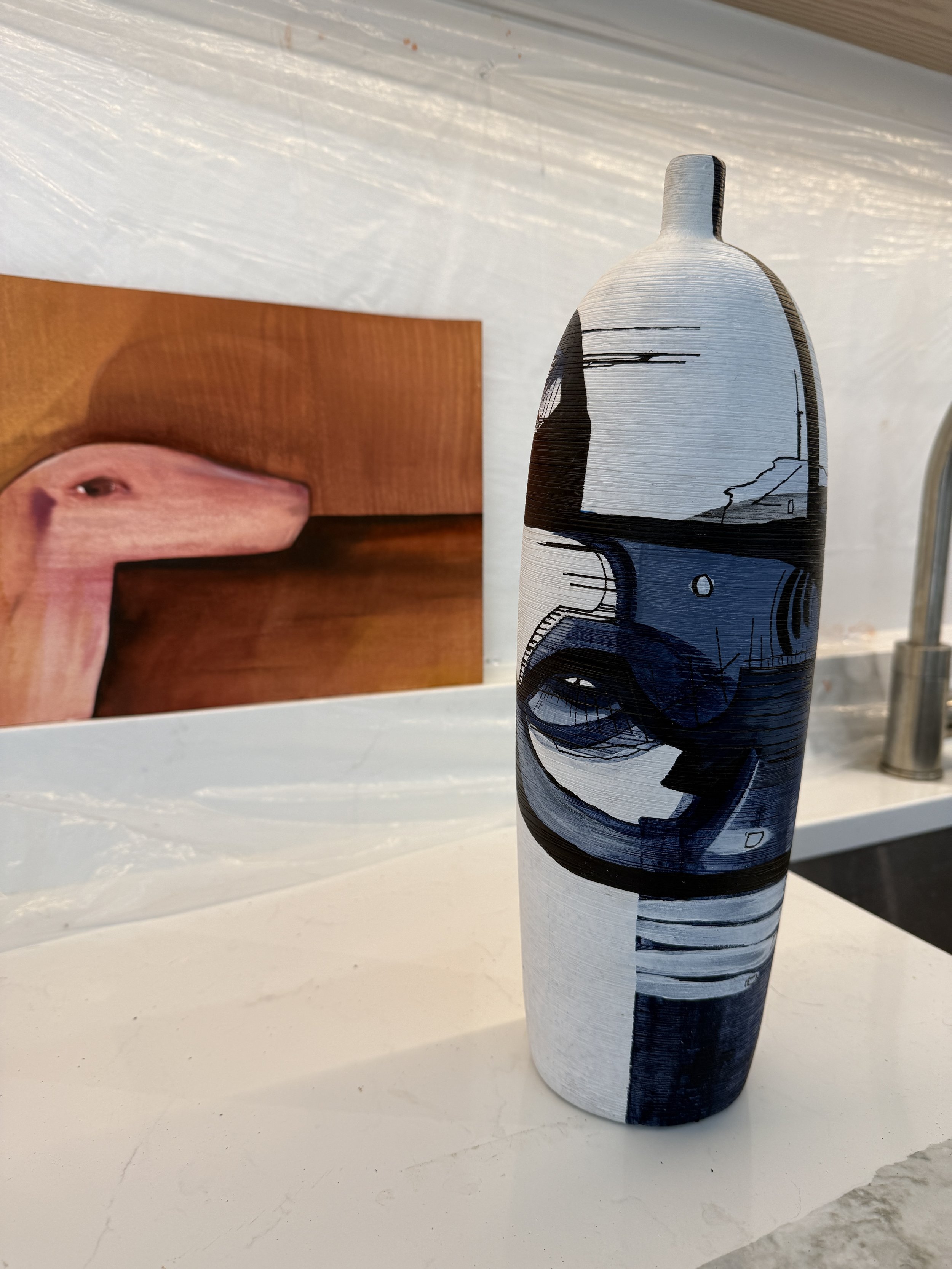 A tall, narrow decorative vase with an abstract black, gray, and white design, placed on a white surface in front of a wooden shelf or panel.