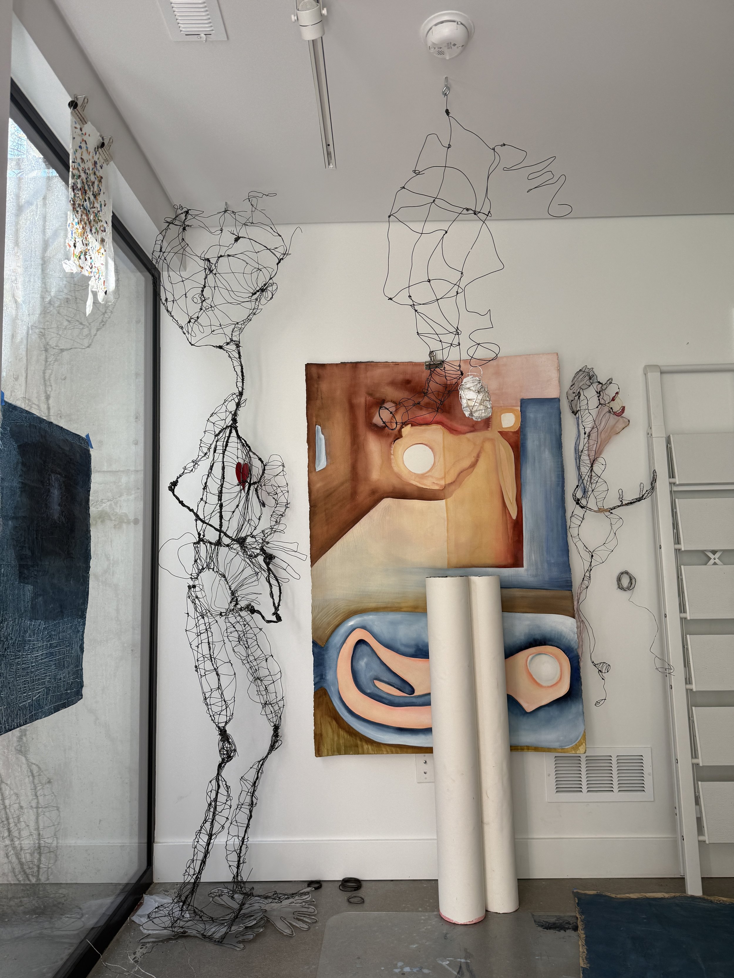 Interior view of an art studio with wire sculptures resembling abstract human forms hanging from the ceiling, a large colorful abstract painting on the wall, rolls of paper or canvas on the floor, and a slanted mirror reflecting part of the room.