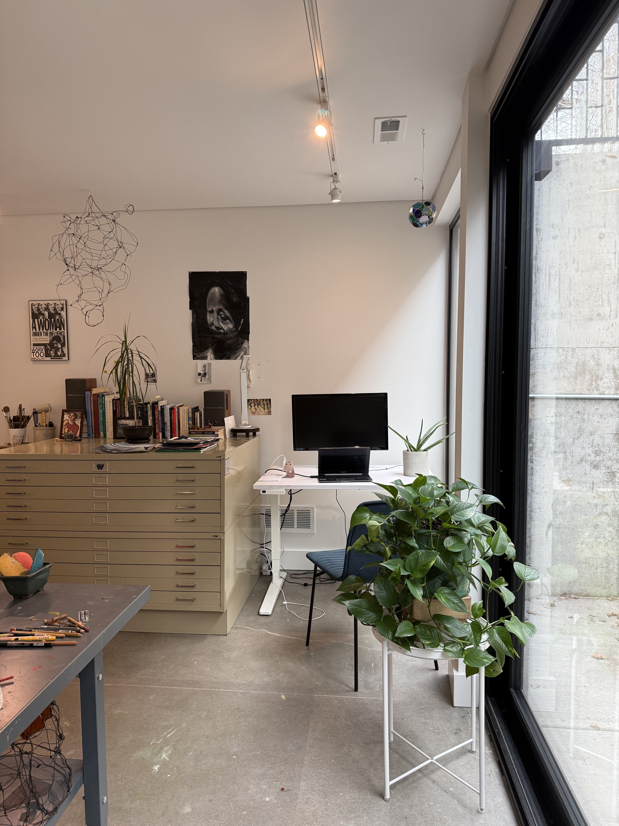 Interior of a modern office or studio space with a large window, green potted plants, a beige filing cabinet, a computer monitor with a small keyboard and mouse, and wall art including a black-and-white face portrait and a poster. The ceiling has tra