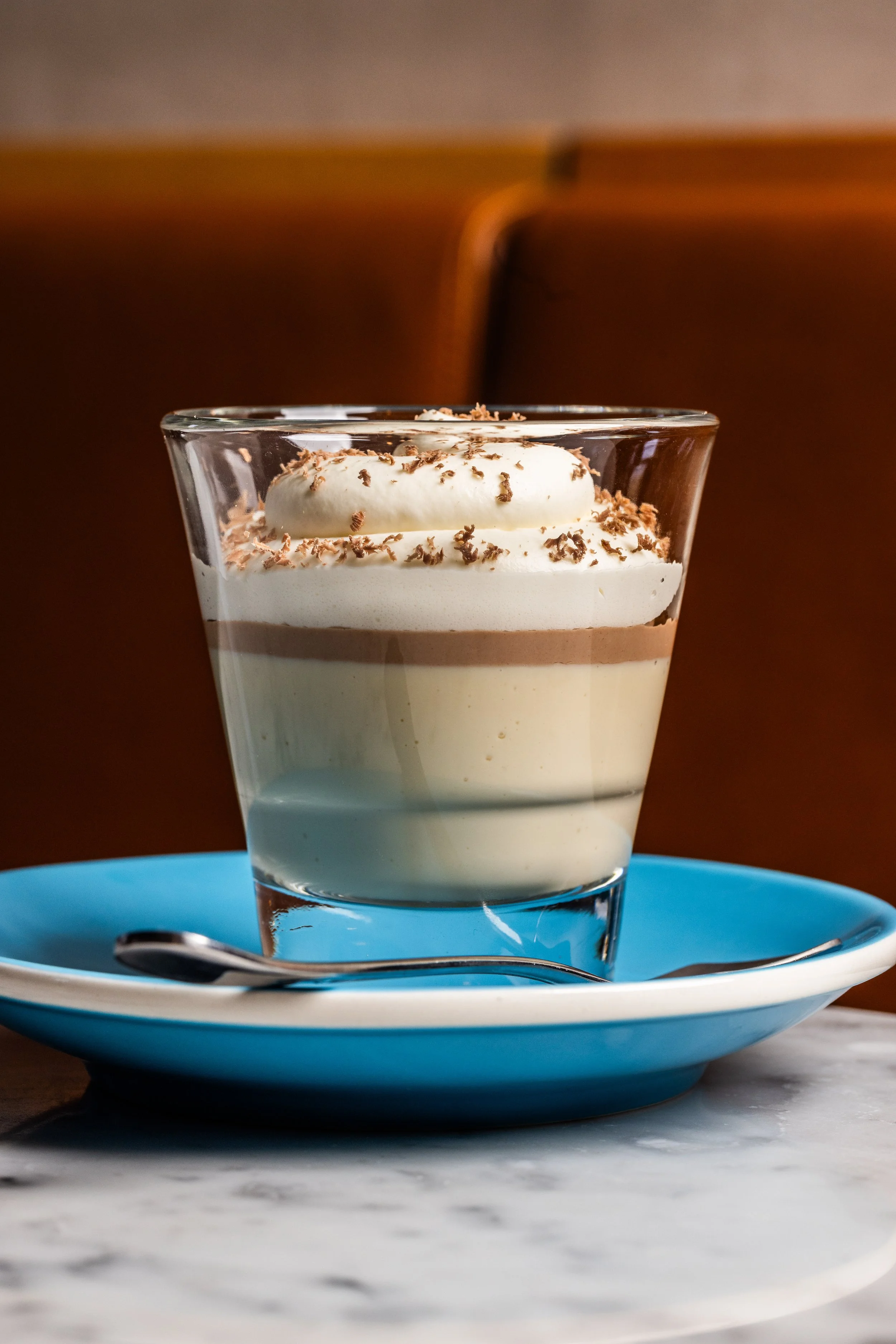 Layered dessert in a clear glass with whipped cream, chocolate shavings, and a spoon on a blue plate.