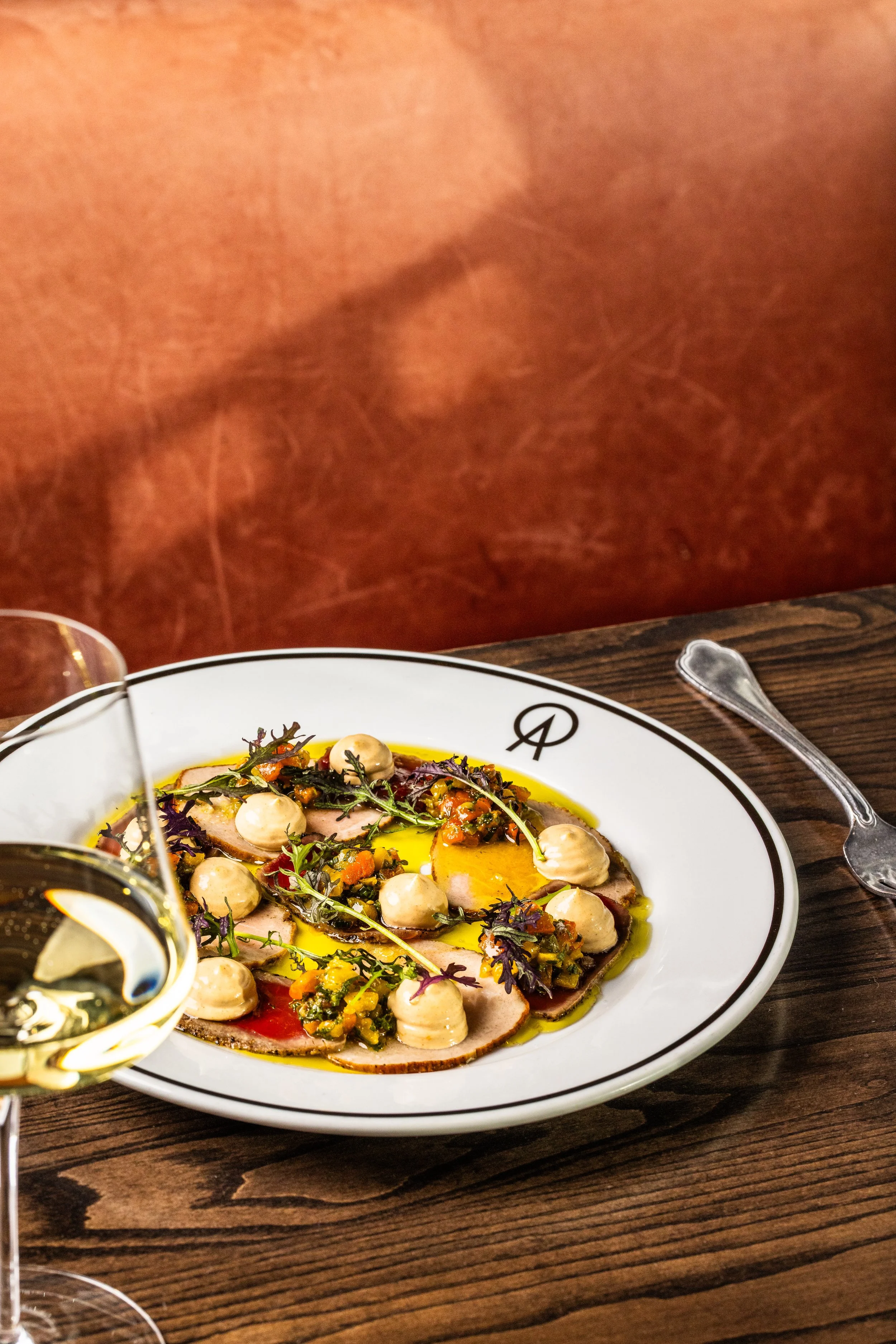 A plate of assorted sliced meats, garnished with herbs, dotted with dollops of a creamy sauce, and drizzled with olive oil, served on a white plate with a black outline and logo.