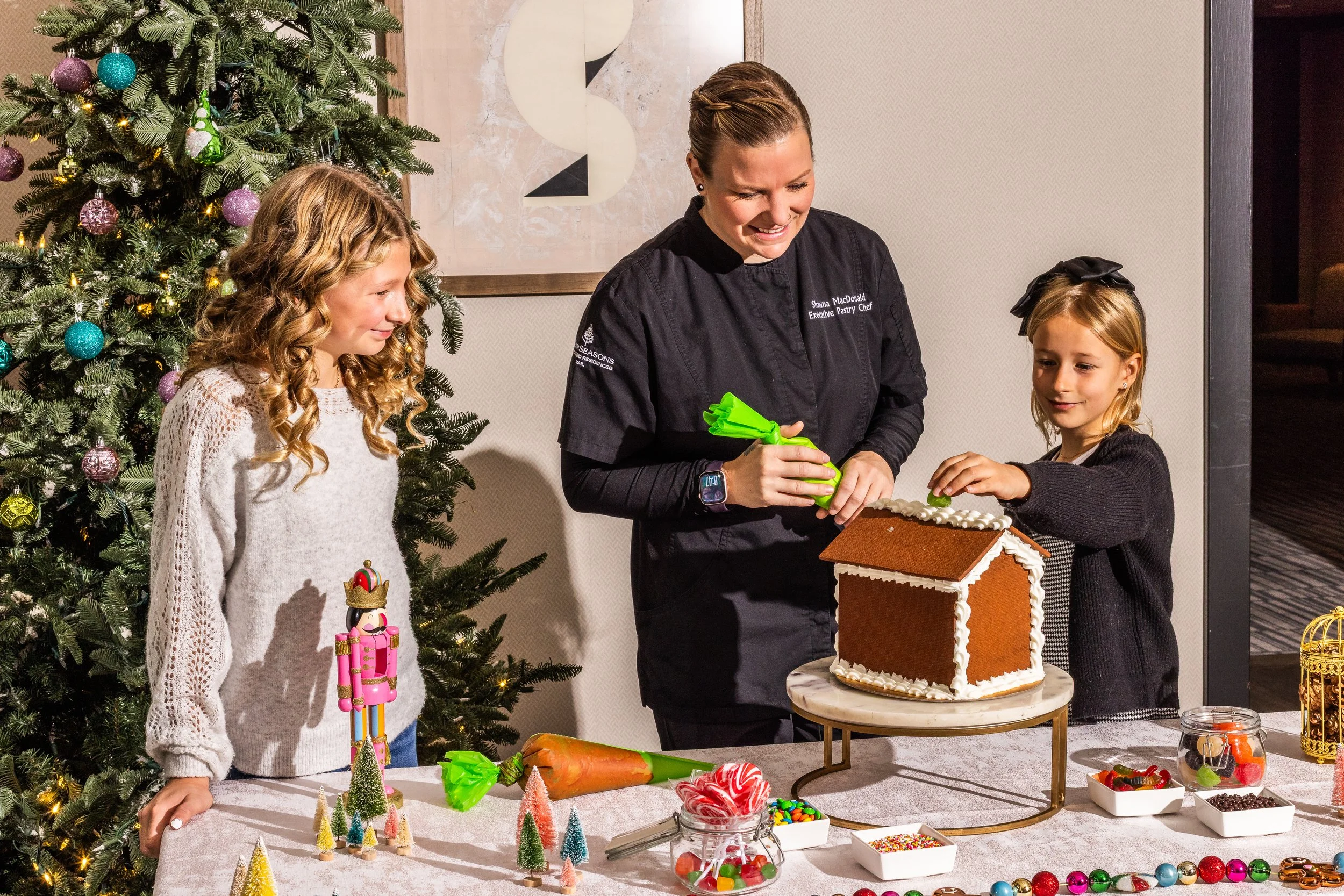 A woman and two young girls decorating a gingerbread house at a holiday gathering. The woman is holding a piping bag, and the girl on the right is placing a decoration on the house. There is a decorated Christmas tree and holiday treats on the table.