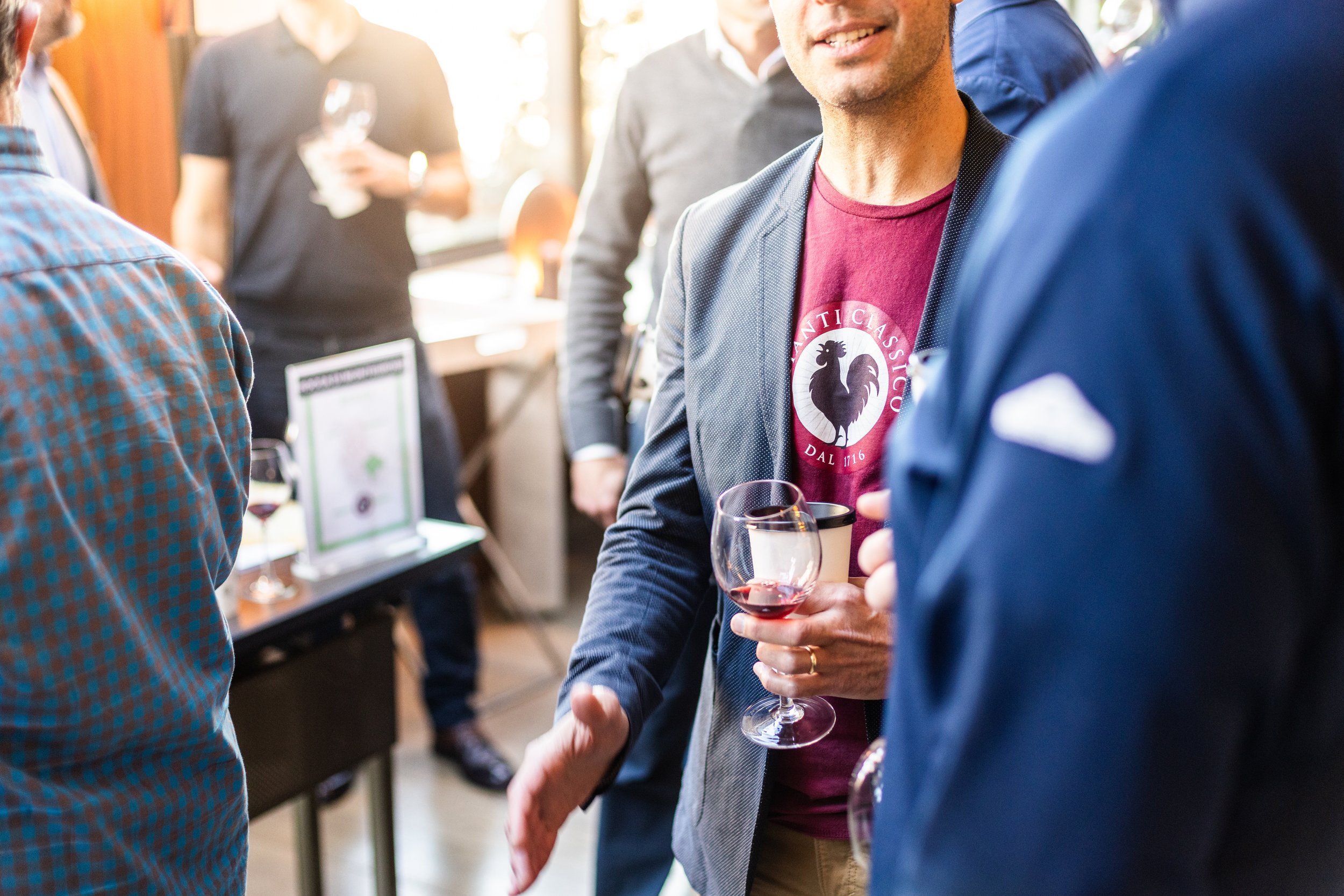 People socializing at a wine tasting event, holding glasses of red wine, in a well-lit indoor space.