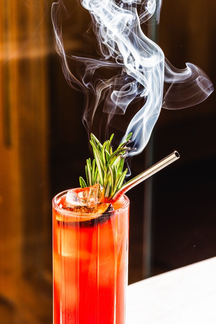 A tall glass with a red drink, garnished with rosemary and ice, emitting smoke with a metal straw.