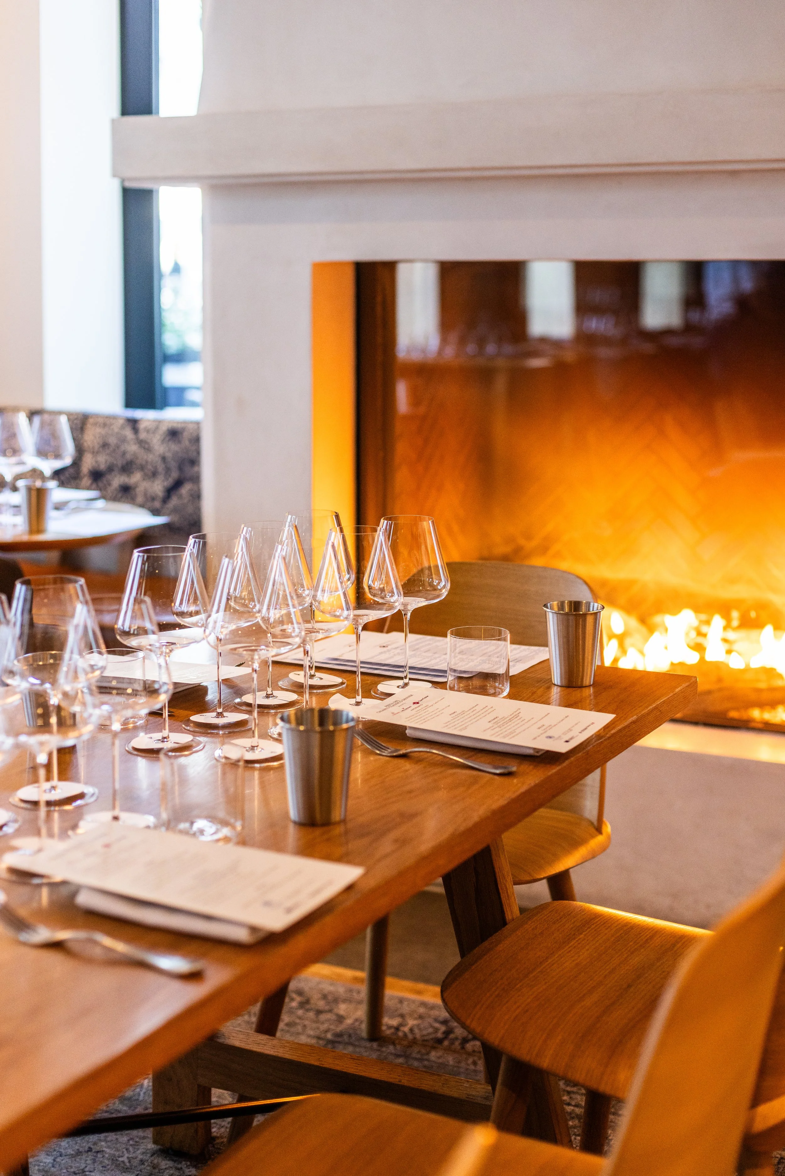 A set dining table in a restaurant with wine glasses, menus, and metal cups, near a fireplace with orange flames.
