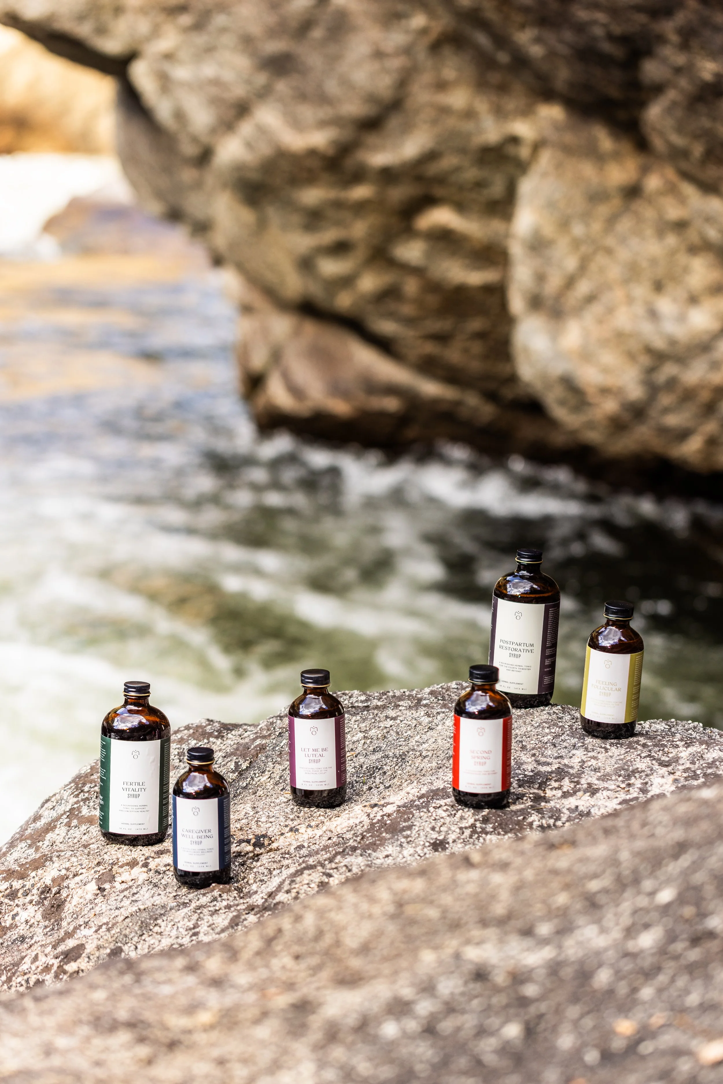 Seven bottles of skincare or wellness products placed on a large rock by the water at a natural outdoor setting.