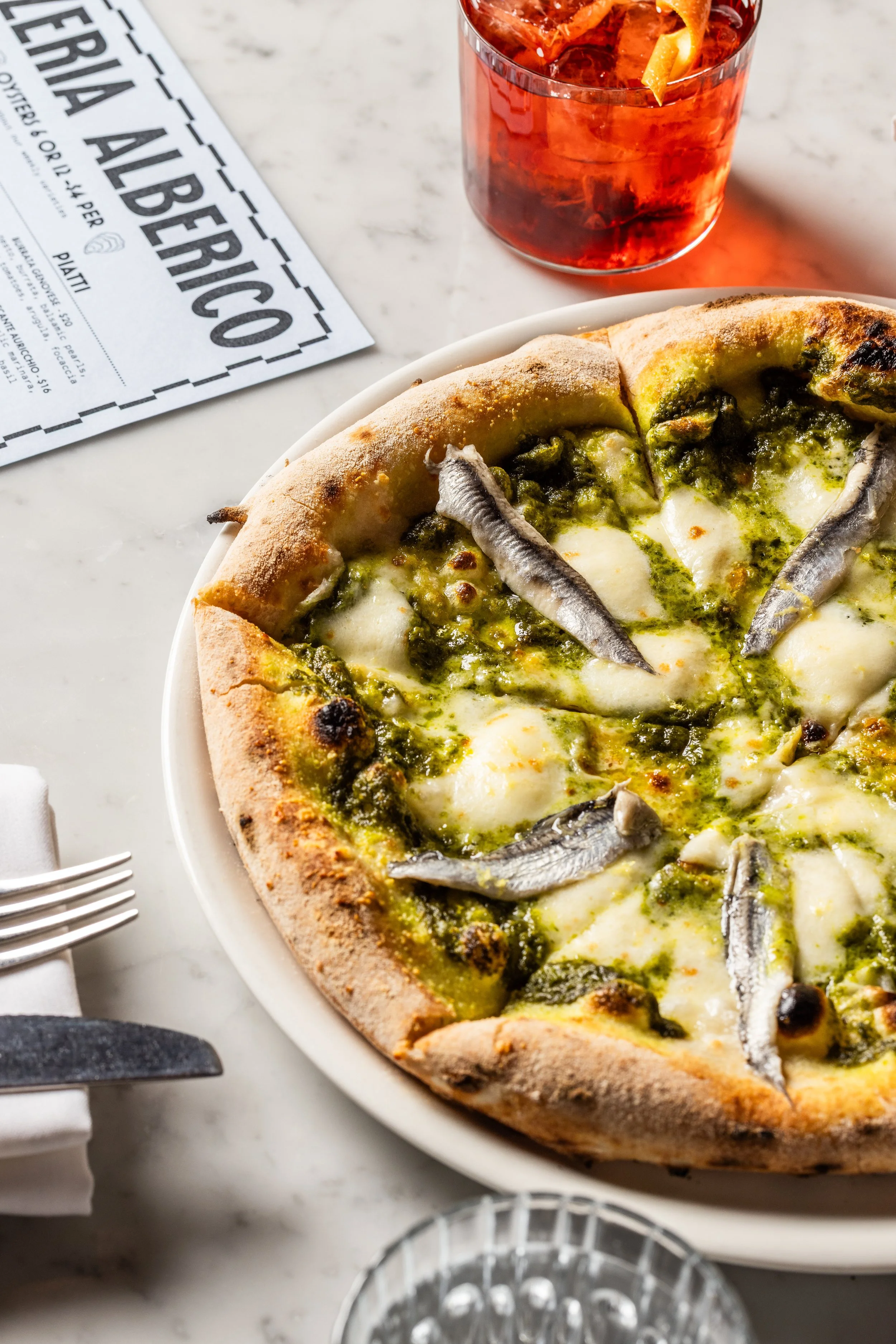 A pizza with cheese, green pesto, and anchovies on a white plate, with a glass of red-colored drink and a menu paper on a white marble table.