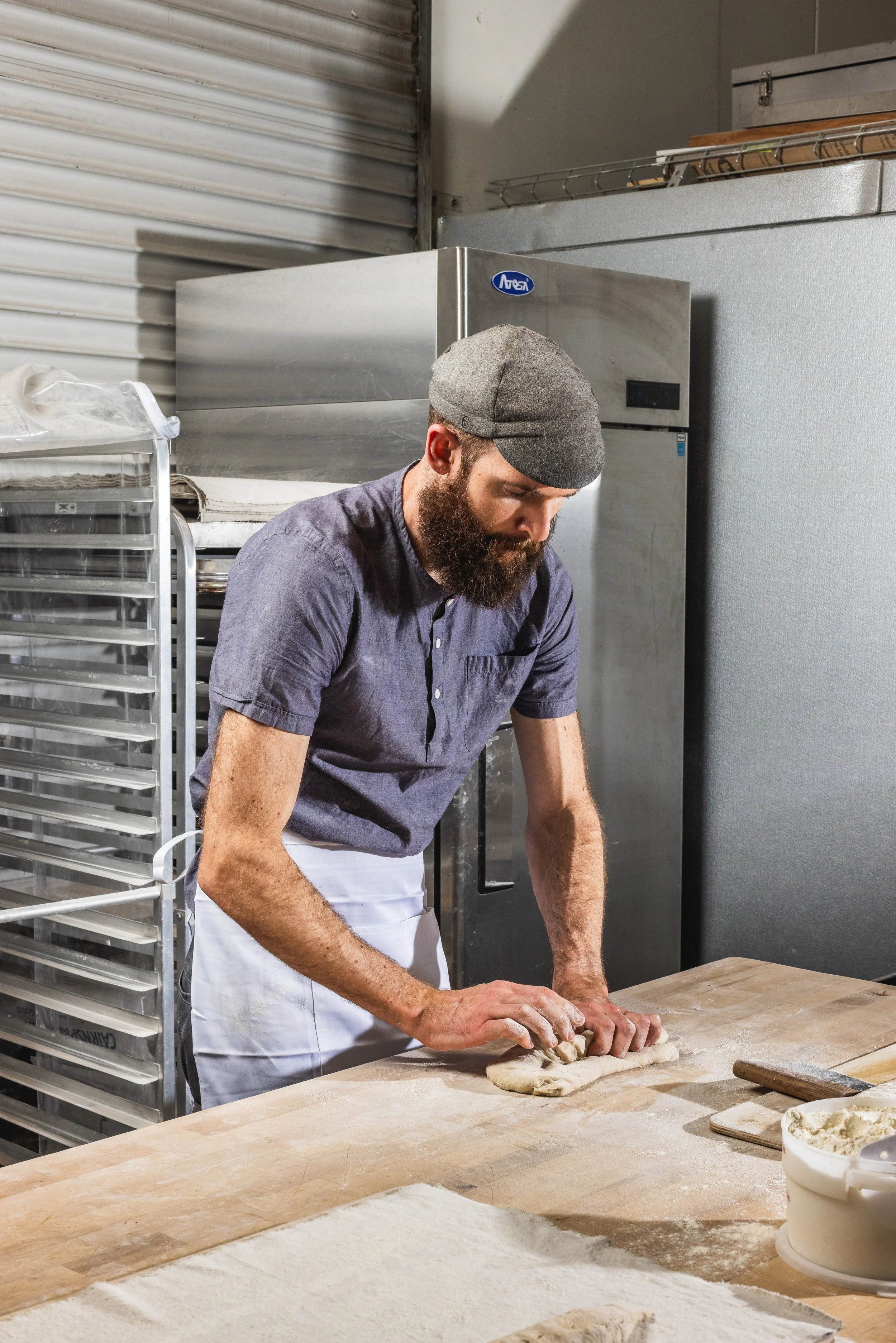 A man with a beard, wearing a gray cap and blue shirt, is kneading dough on a wooden work surface in a bakery kitchen.