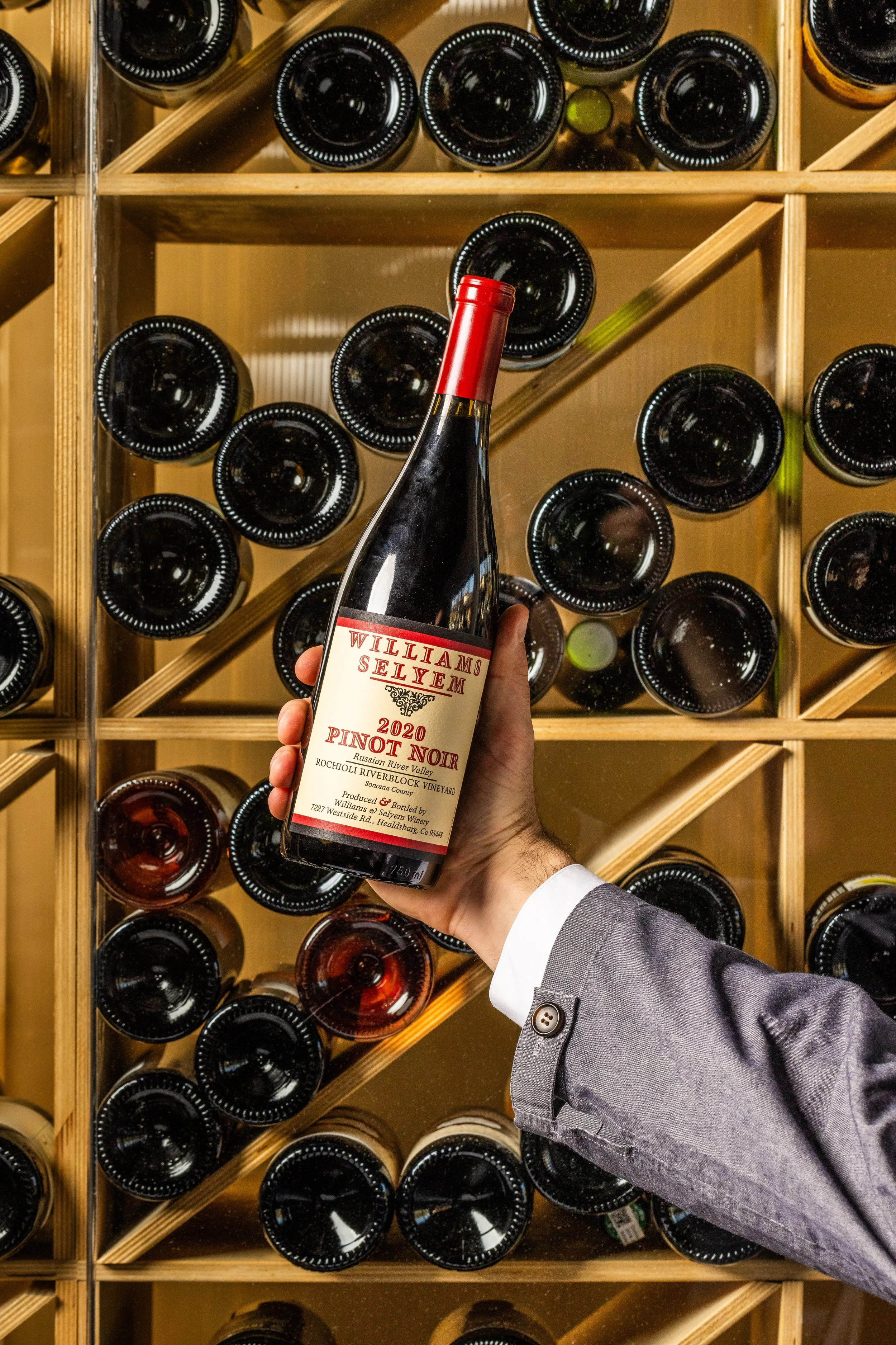 Hand holding a bottle of Williams Selyem Pinot Noir wine in front of a wine rack filled with bottles.