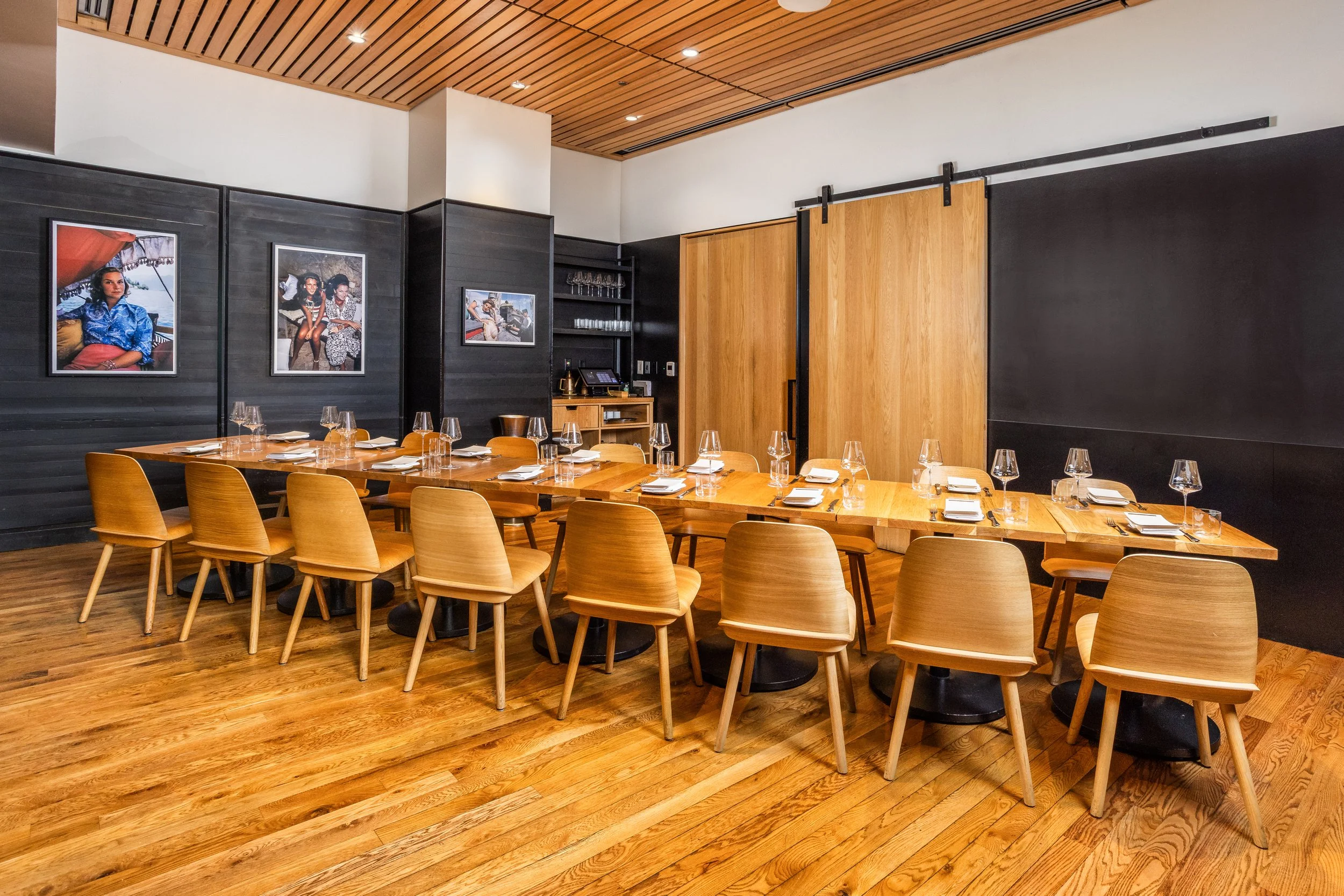 Empty restaurant dining area with wooden chairs and a large wooden table set with glasses, plates, and napkins. The wall features framed photos, and there is a wooden sliding door in the background.