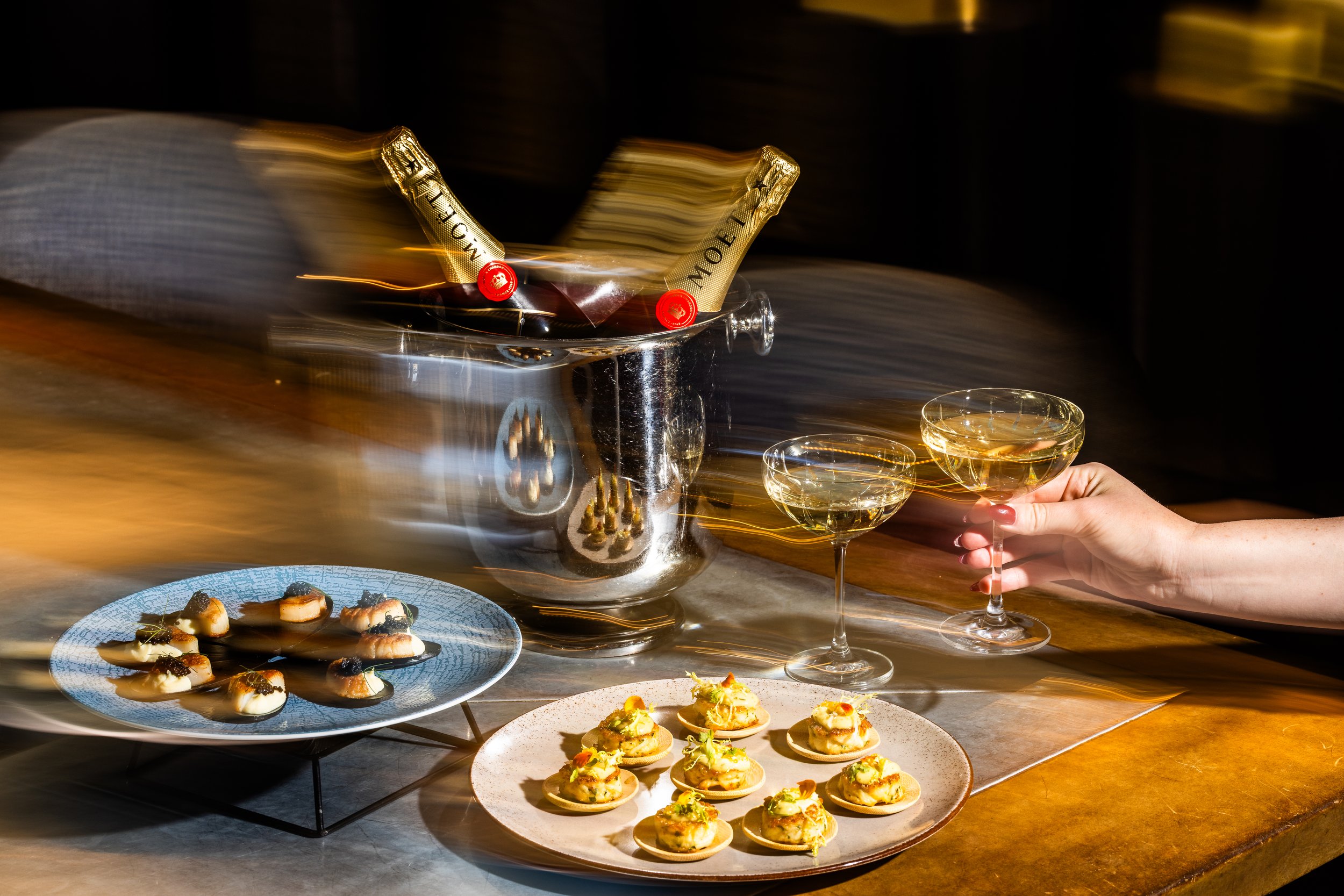 Two glasses of white wine or champagne held by a woman's hand, a silver ice bucket with bottle of Moët Champagne, two plates of appetizers on a wooden table, with a breakfast-style dish with small toppings, all in a dimly lit setting.