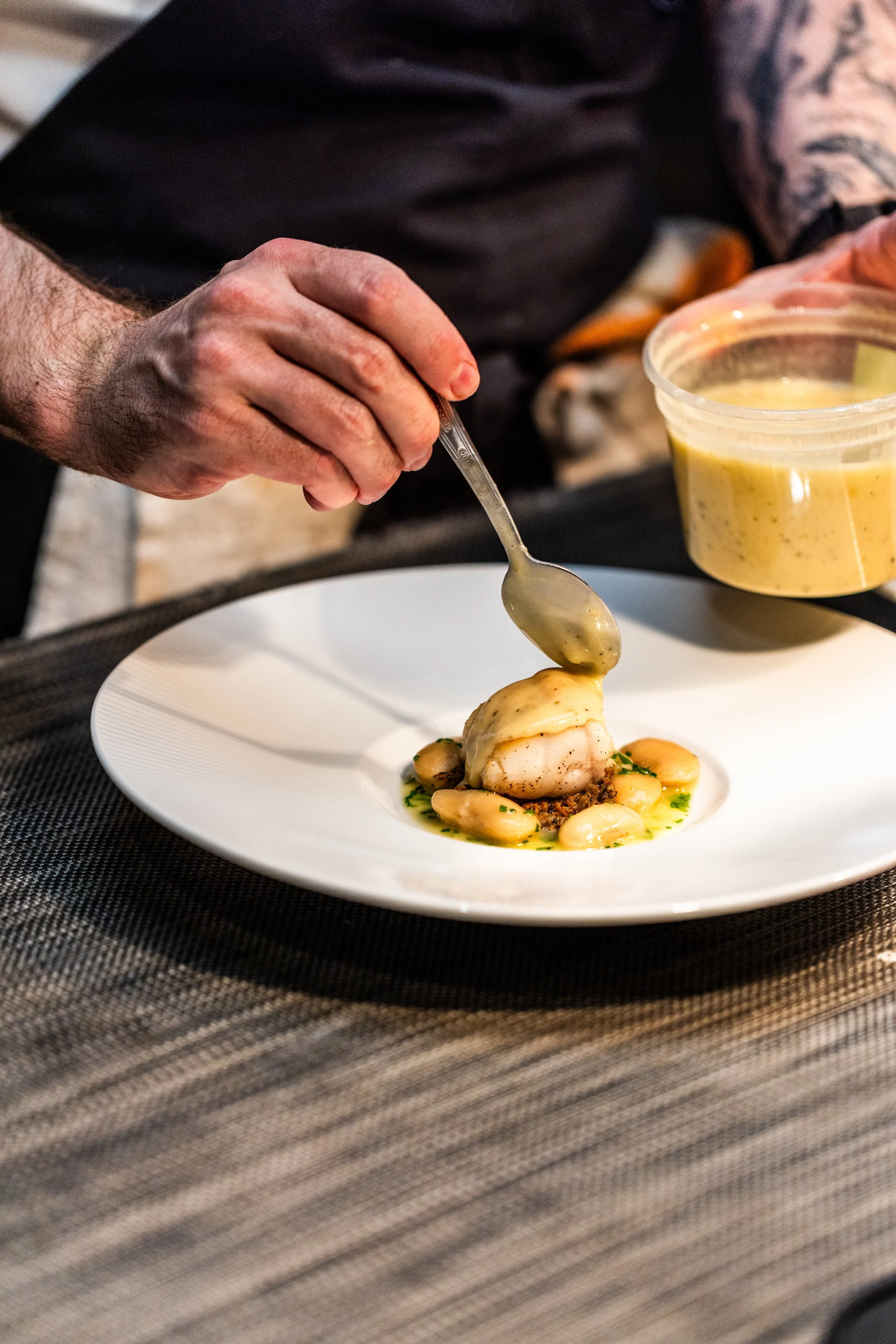 A chef drizzling sauce over a plated gourmet seafood dish with beans and herbs on a white plate.