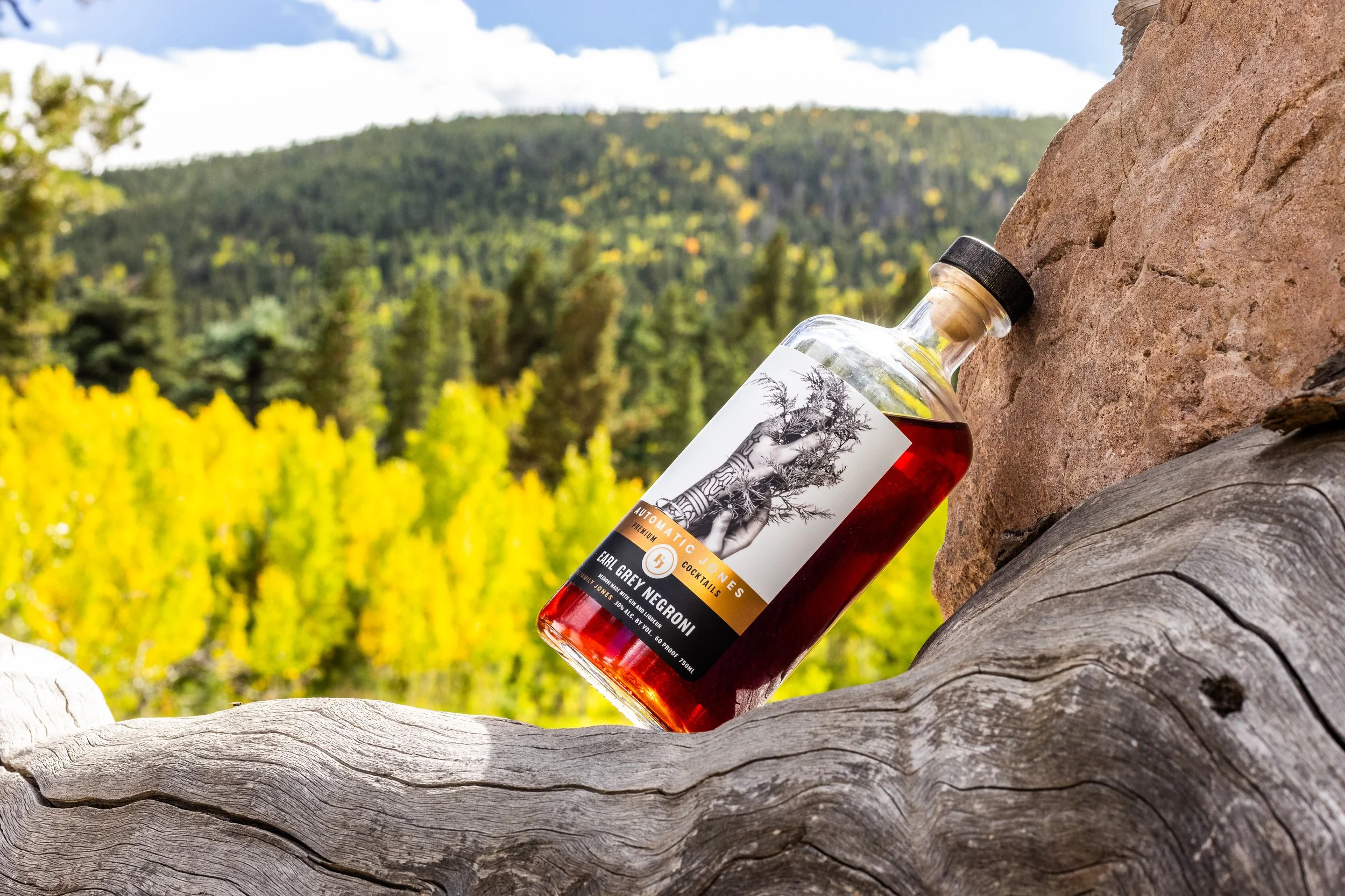 A bottle of Earl Grey Negroni cocktail resting on a weathered wooden surface outdoors, with a background of green and yellow foliage and a distant hillside under a partly cloudy sky.