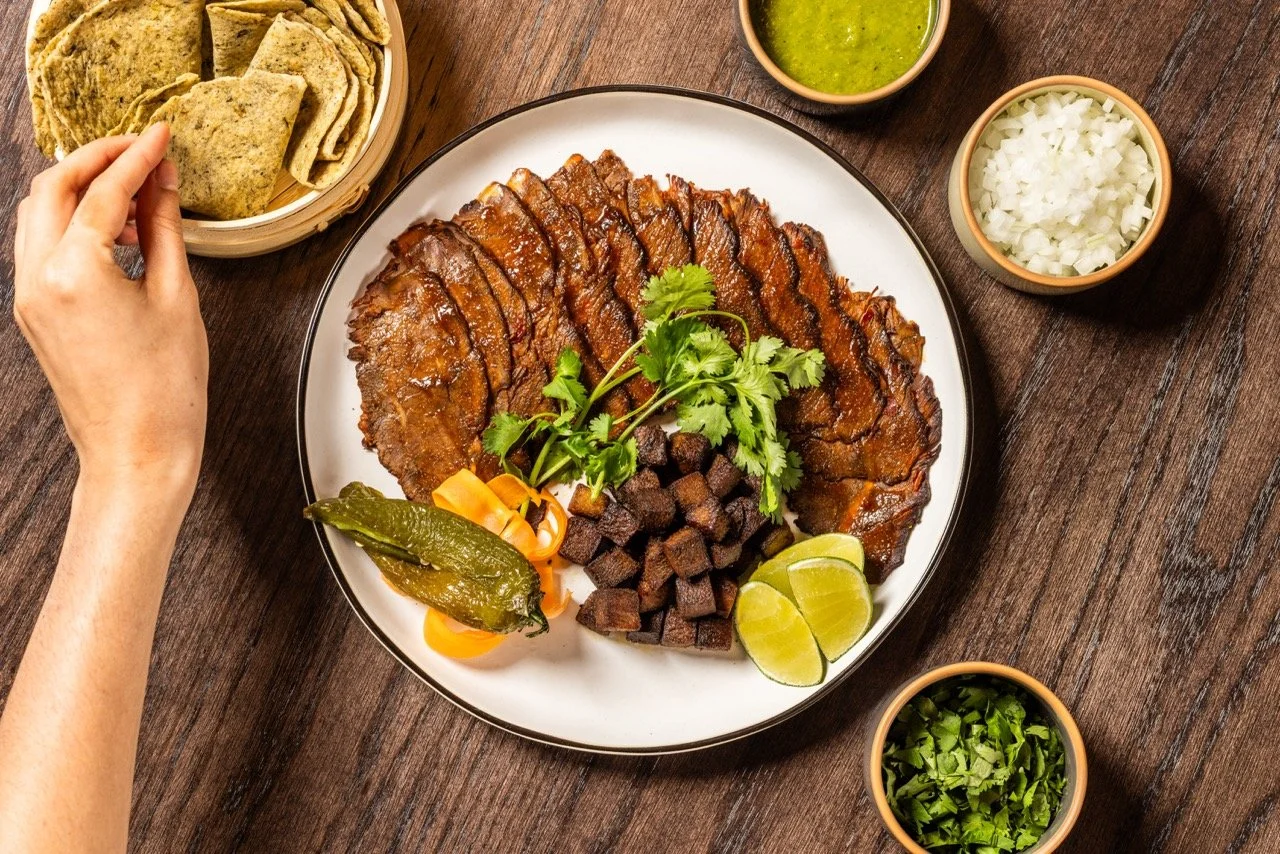 A plate of sliced roasted beef garnished with cilantro, lime wedges, and diced meat, accompanied by bowls of chopped onions, salsa verde, chopped cilantro, and corn tortillas, on a wooden table.
