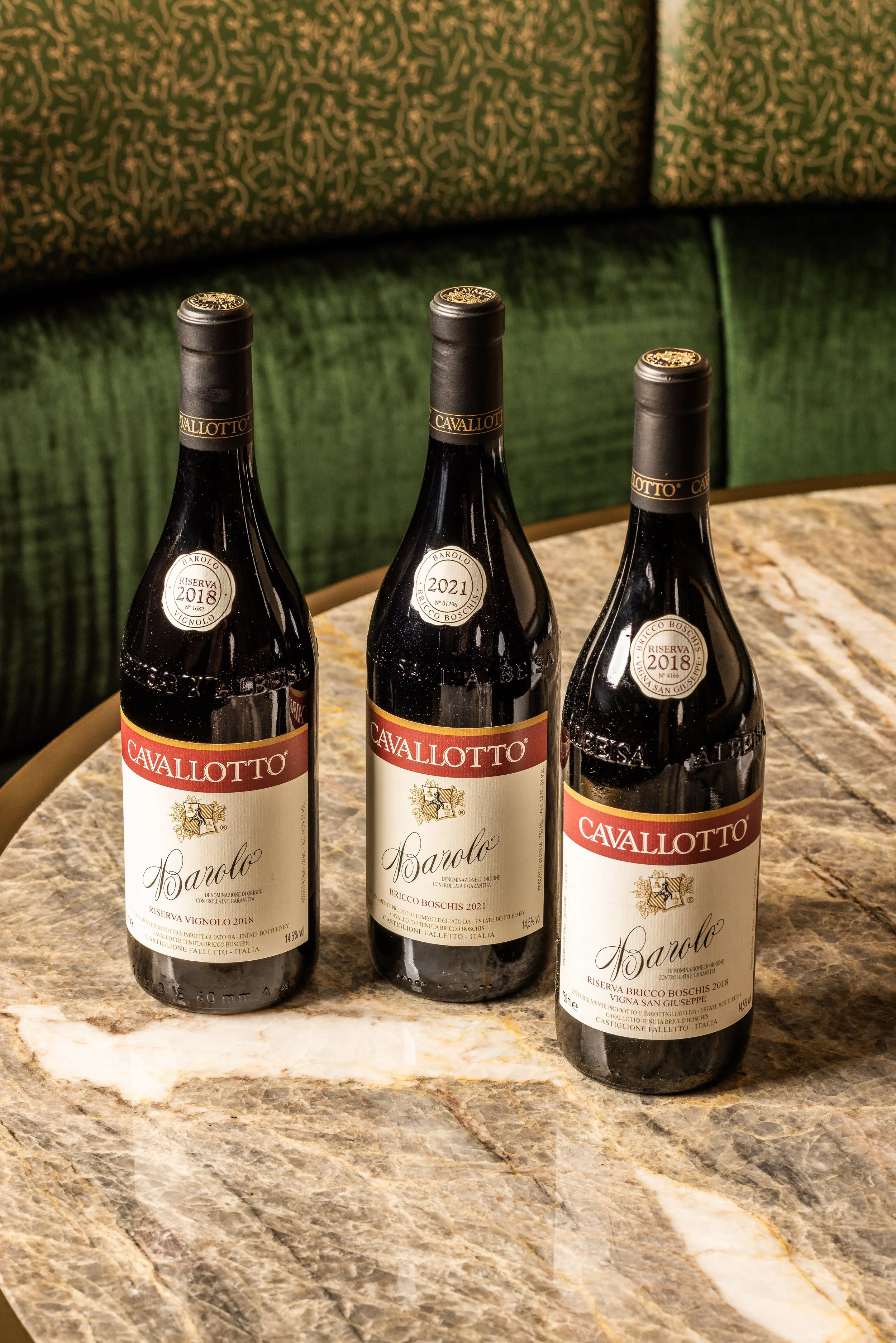 Three bottles of Cavallotto Barolo wine on a marble table in front of green upholstered chairs.